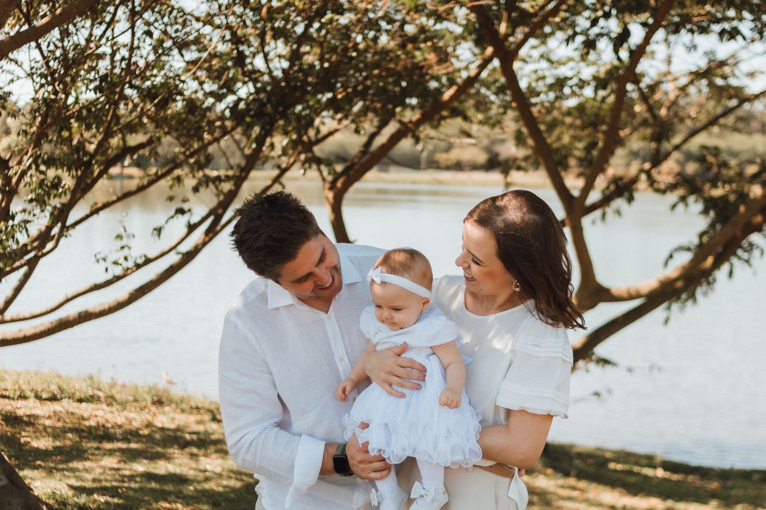 A happy family in white outfits holds their baby girl outdoors near a lake under a tree.