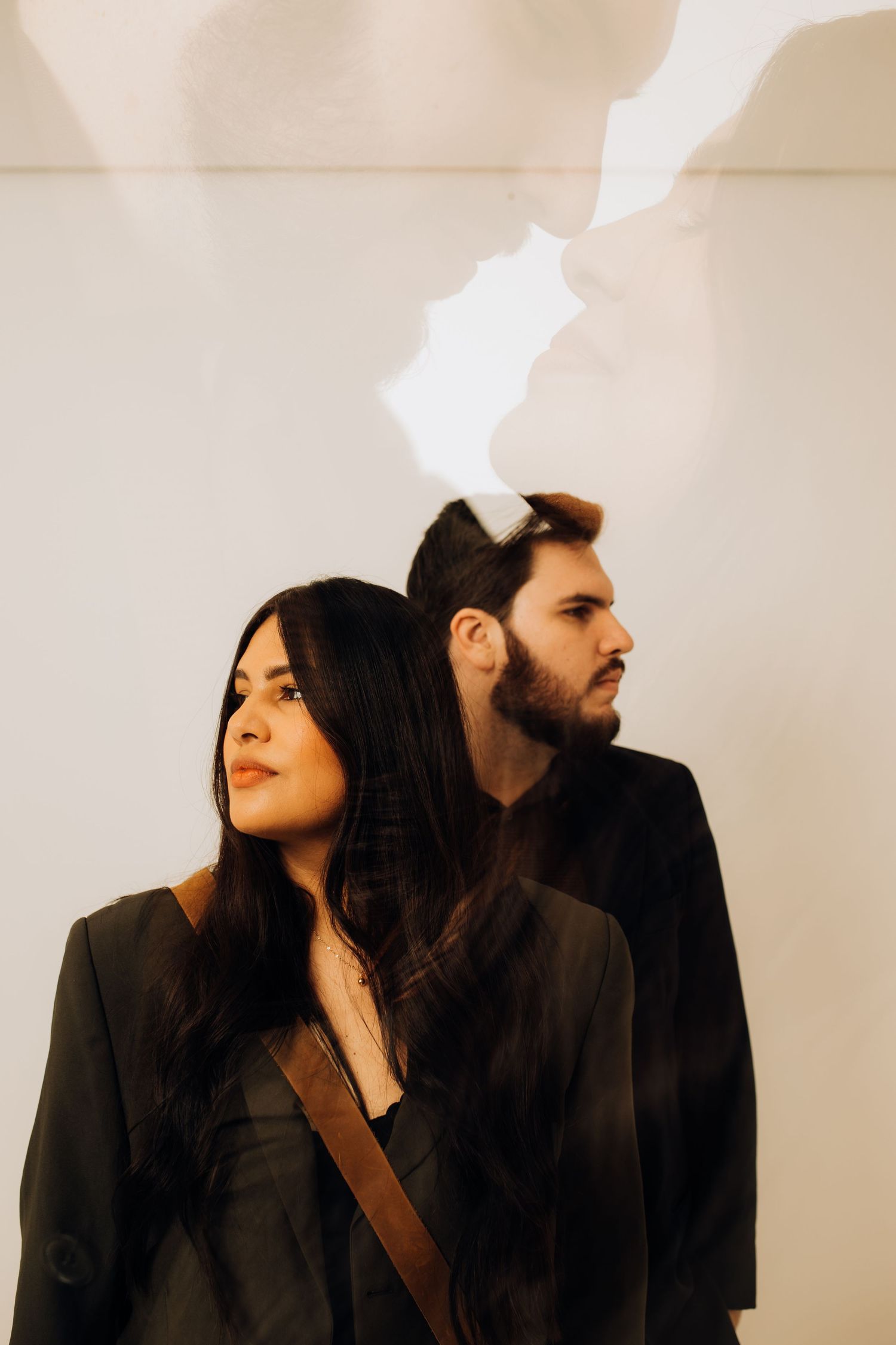 A woman and man in dark blazers stand before a white wall with an artistic light projection silhouette above them.