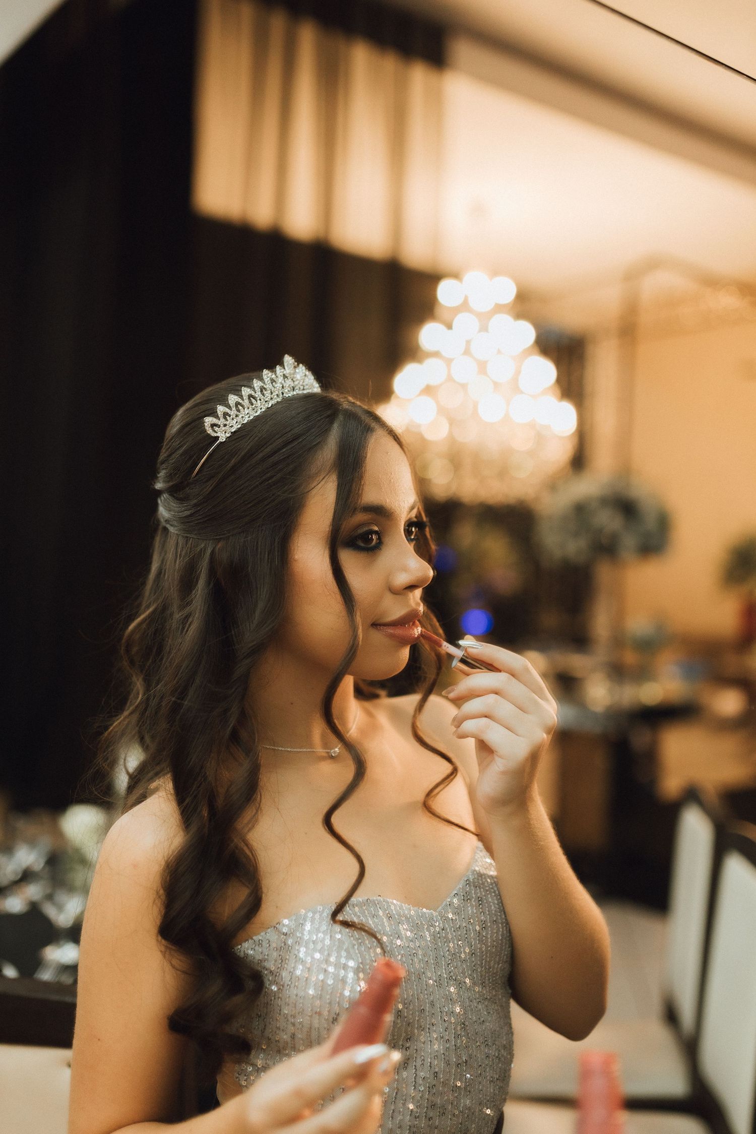Young woman in a sparkling silver gown and tiara applies lipstick while preparing for a formal event.
