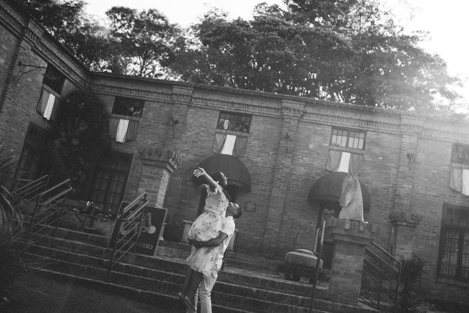 Black and white photo of a person standing on steps before a historic brick building surrounded by trees.