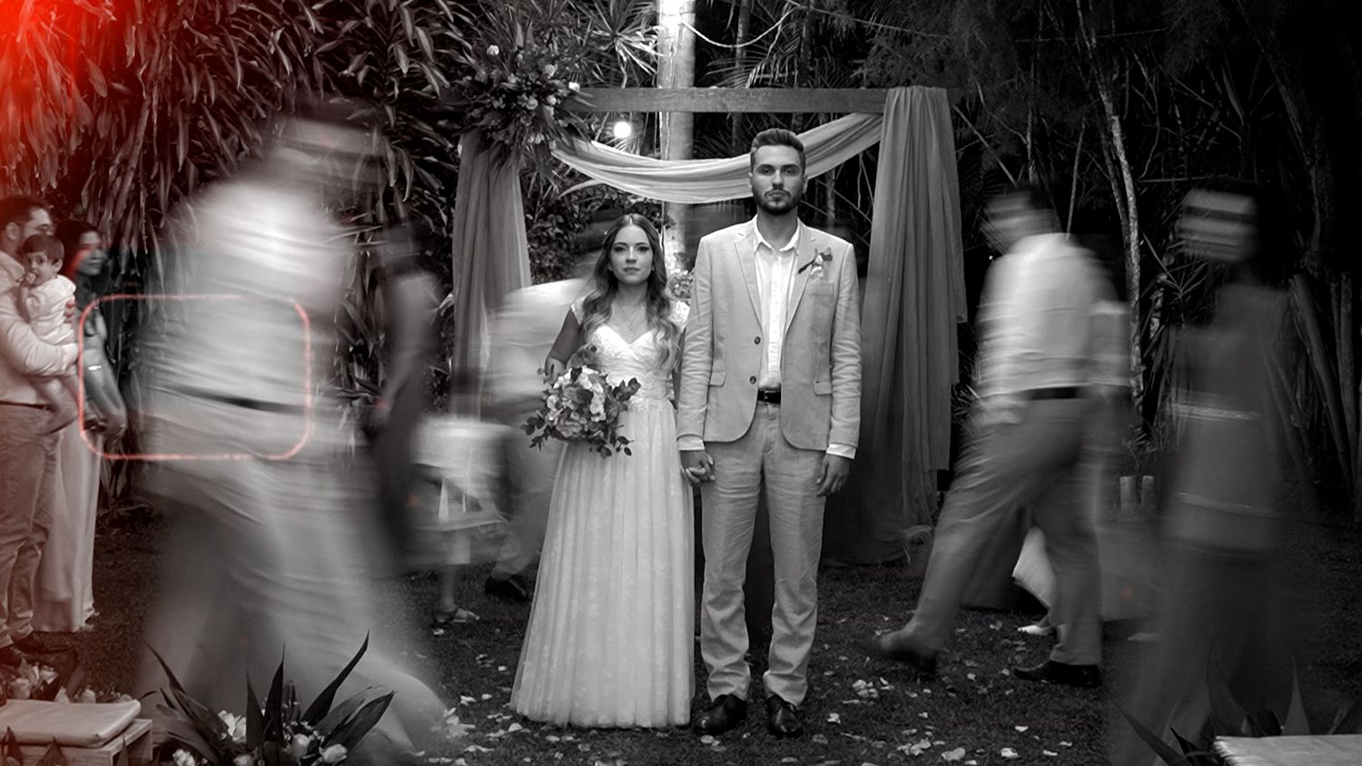 Black and white wedding photo of bride and groom standing still amid blurred moving guests near a cross.