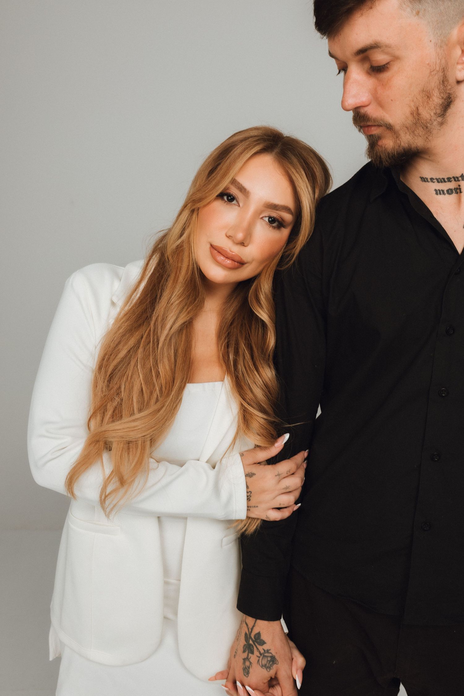A blonde woman in a white blazer leans against a tattooed man in a black shirt against a gray background.