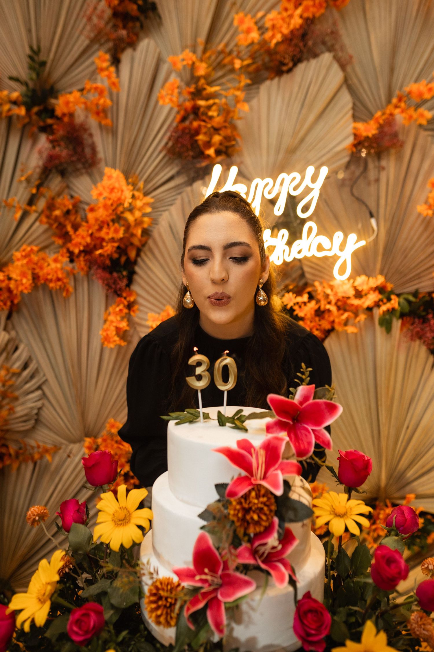 Woman blowing out 30th birthday candles on white floral cake with Happy Birthday neon sign backdrop.