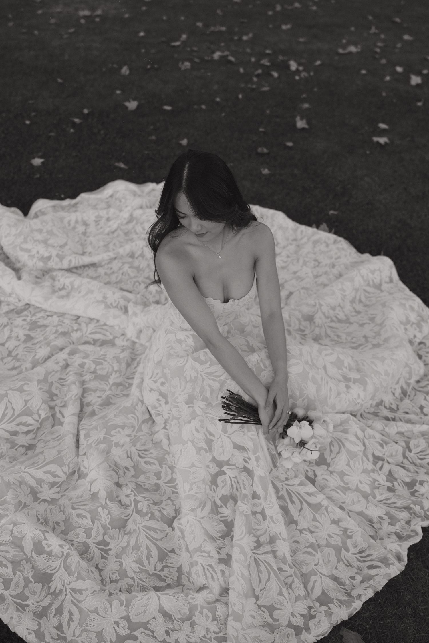 Black and white photo of a bride sitting on grass holding a bouquet on her wedding day.