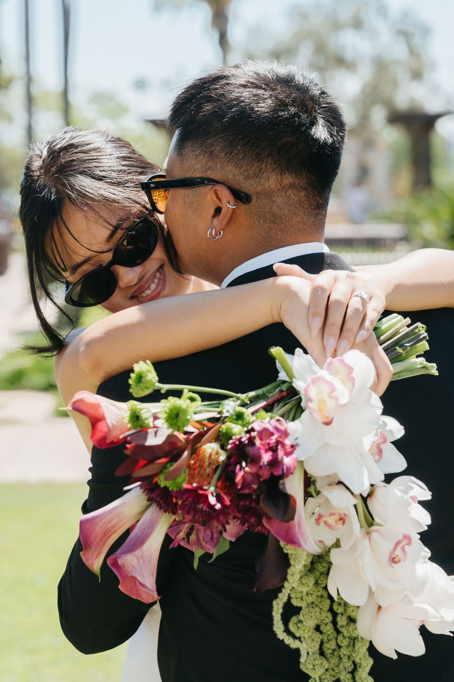 Bride and groom embracing outdoors, bride smiling in sunglasses holding a colorful floral bouquet on a sunny day during their elopement.