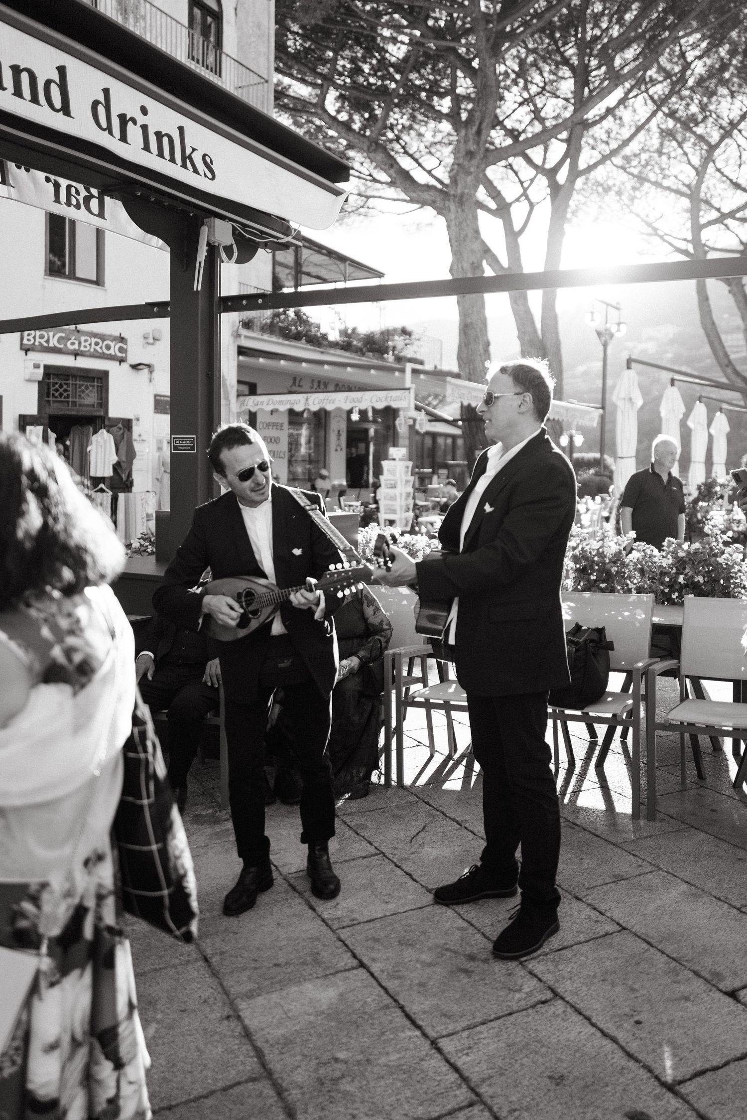 Two men in suits perform outdoors, one playing mandolin, near a cafe in a black and white photo during Cocktail hour on the Amalfi Coast.