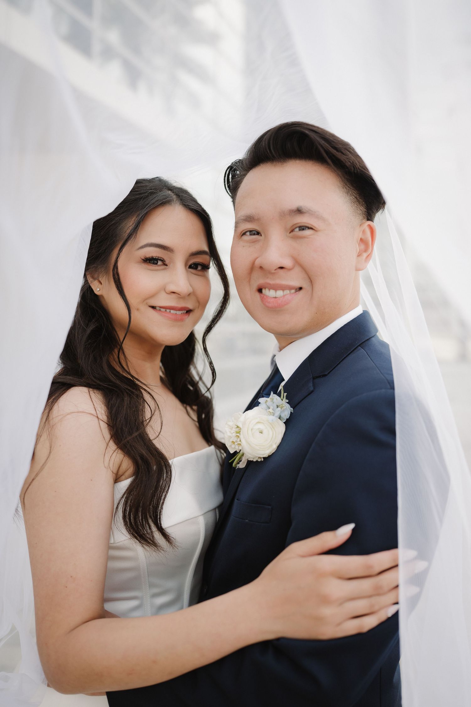 A smiling bride and groom pose together under a white veil, wearing wedding attire with a floral boutonniere.