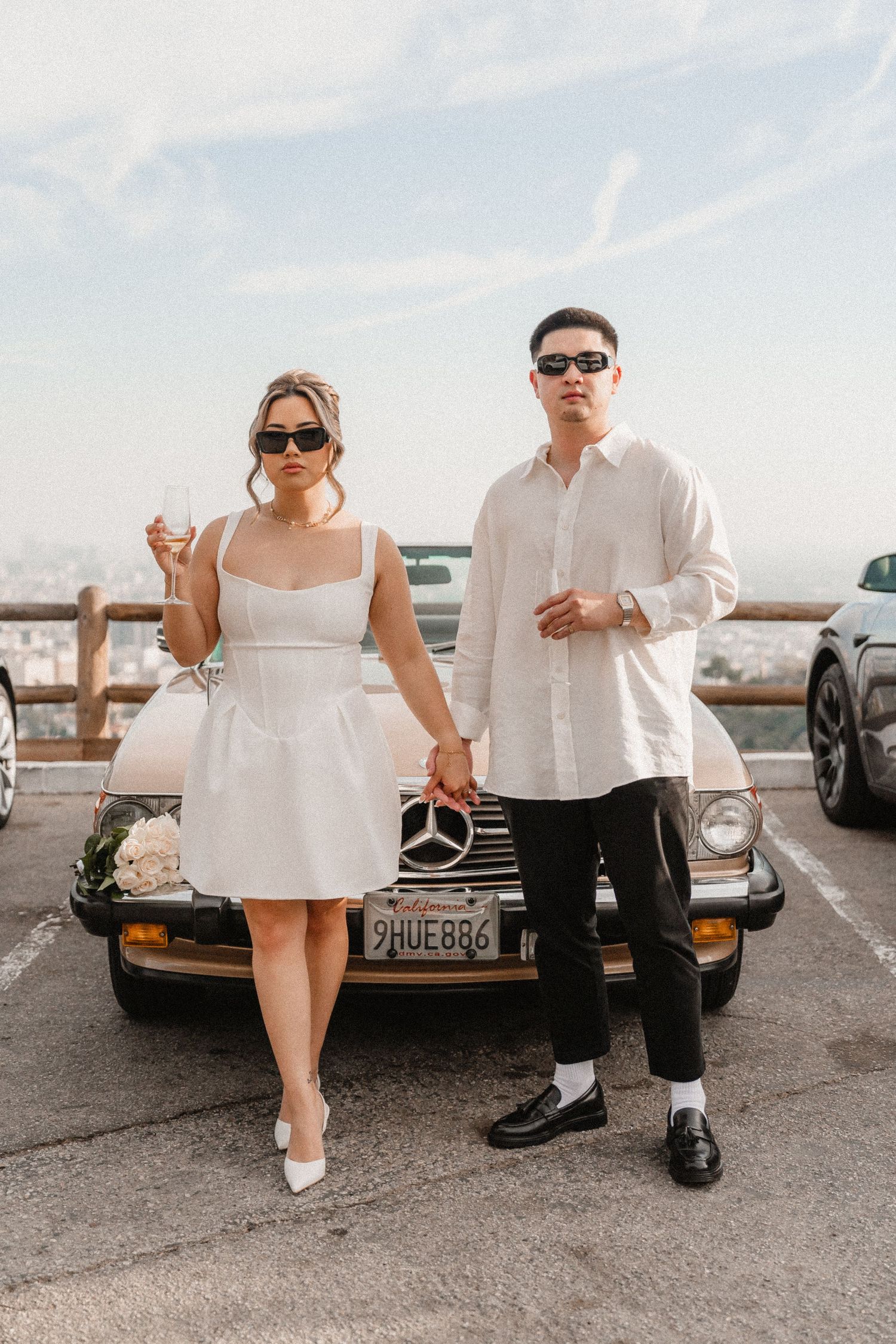 Stylish couple in sunglasses holding hands by a vintage Mercedes convertible, woman in white mini dress, man in white shirt.