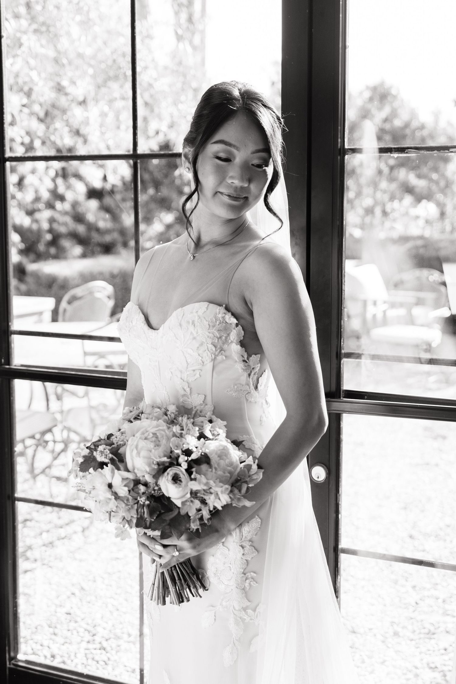 Elegant bride in lace gown holding floral bouquet stands by French doors in black and white wedding portrait.