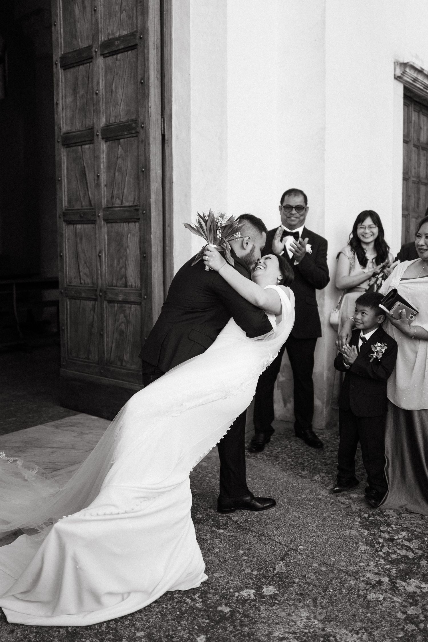Black and white photo of a groom dipping his bride for a kiss outside a church as wedding guests applaud in Ravello, Italy.