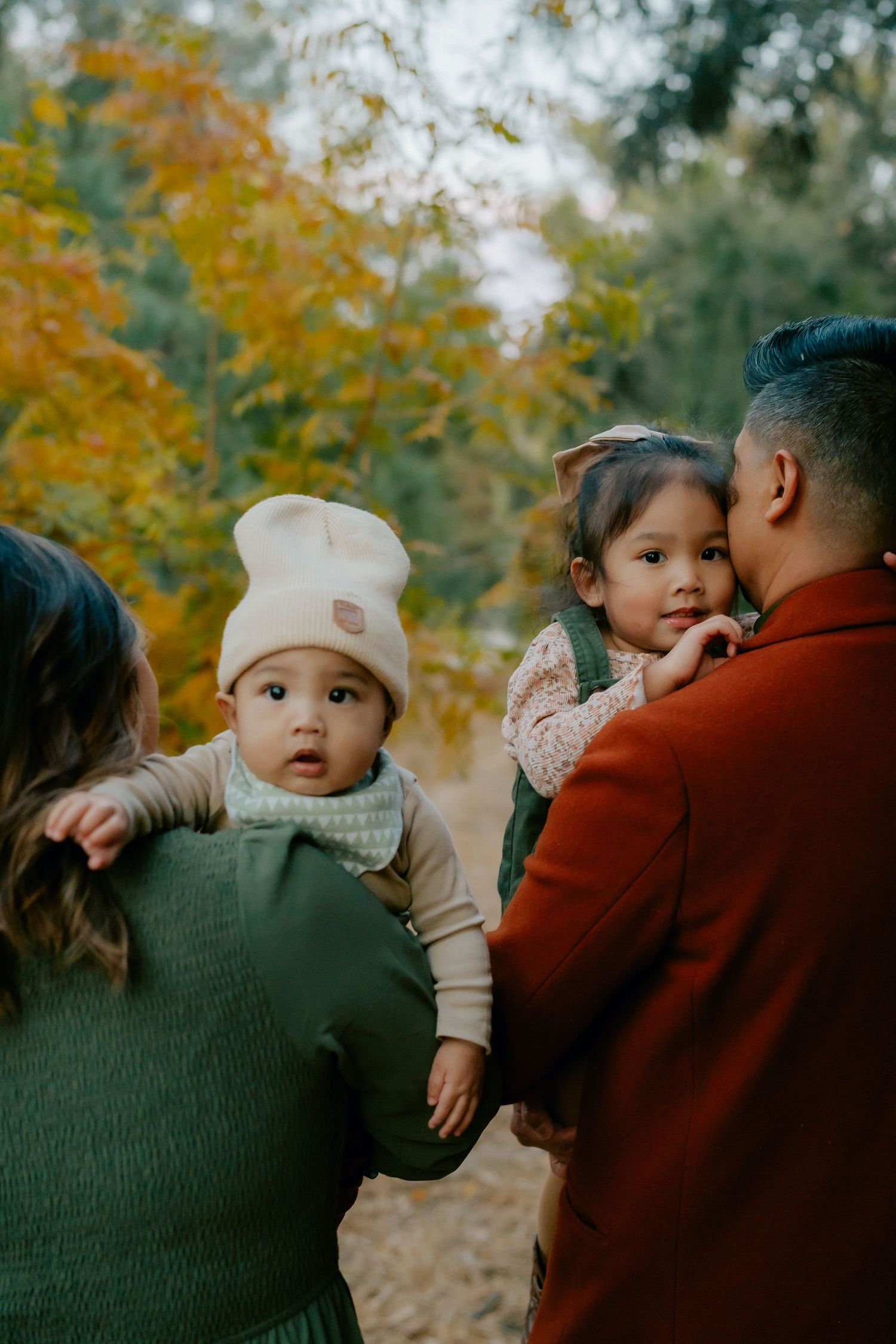 Family with two young children enjoying an autumn outdoor family photoshoot, baby in knit hat and toddler girl held by parents.