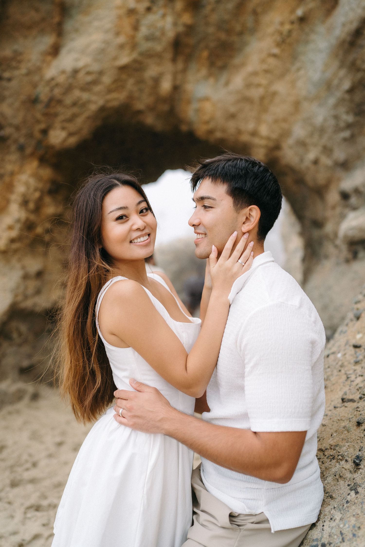 A smiling couple in white outfits embracing romantically against a rocky cliff at Laguna Beach during their engagement session