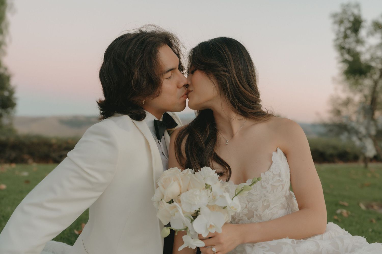 Newlyweds sitting on grass and kissing with a beautiful sunset in the background.