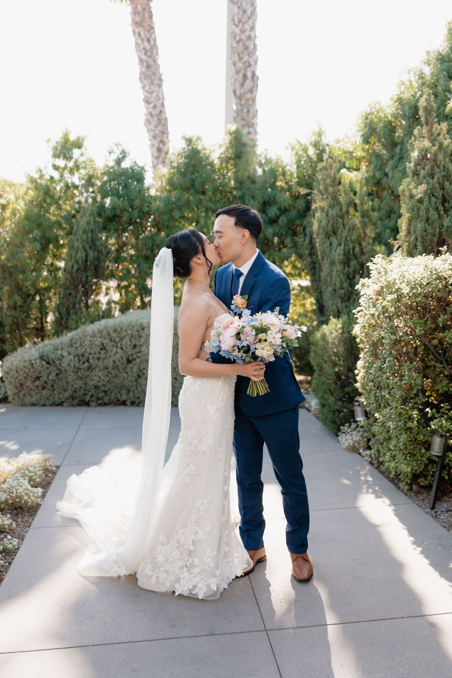 Bride and groom share a kiss on a sunlit garden path, bride in lace gown with veil holding a floral bouquet on their wedding day.