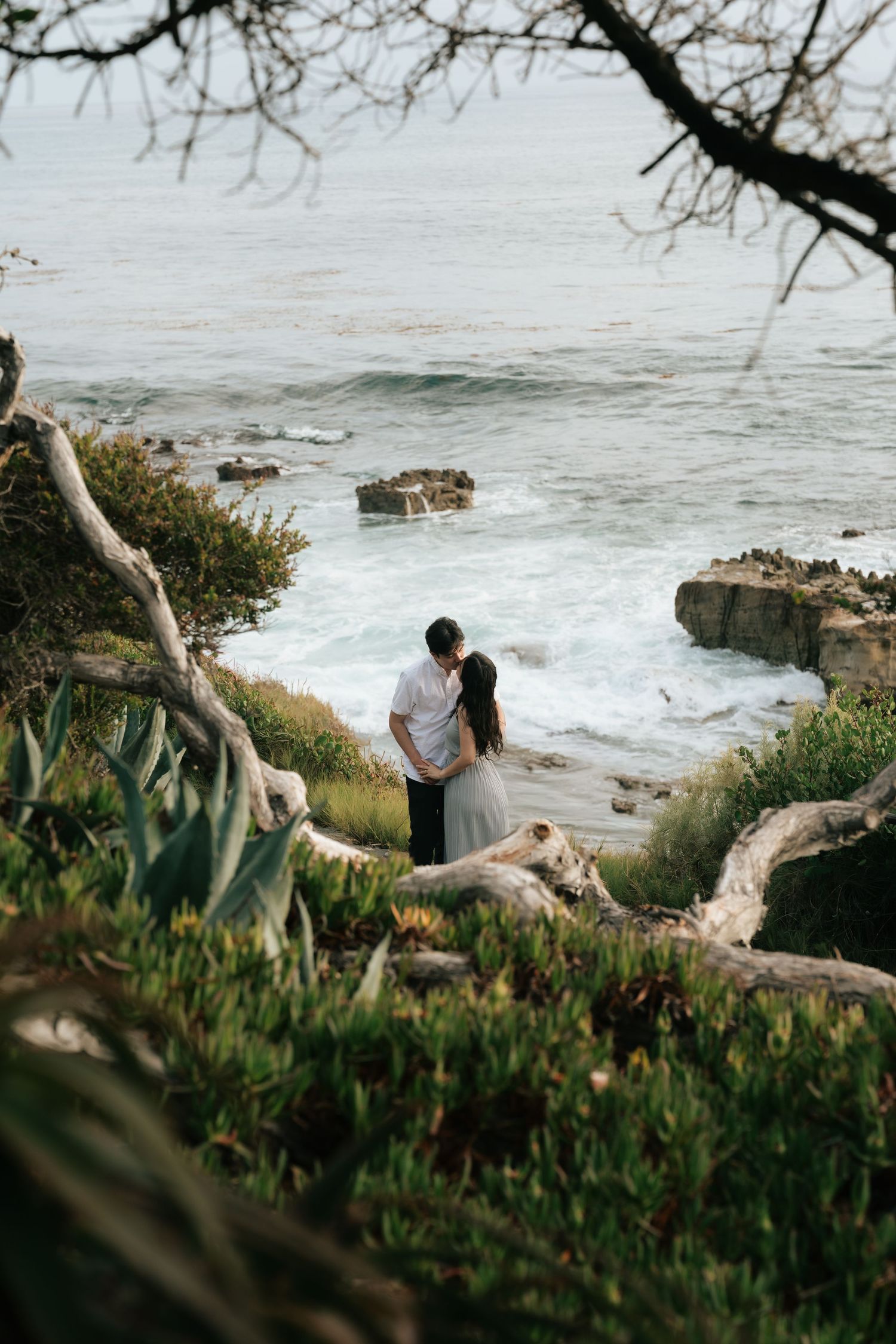 Couple sharing a romantic moment on a coastal cliff surrounded by lush greenery with ocean waves crashing below in Laguna Beach
