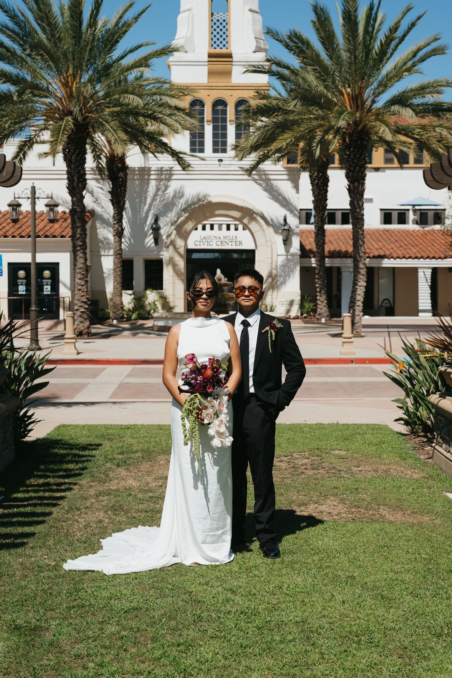 Bride and groom pose in front of a Spanish-style civic center surrounded by palm trees on their wedding day.