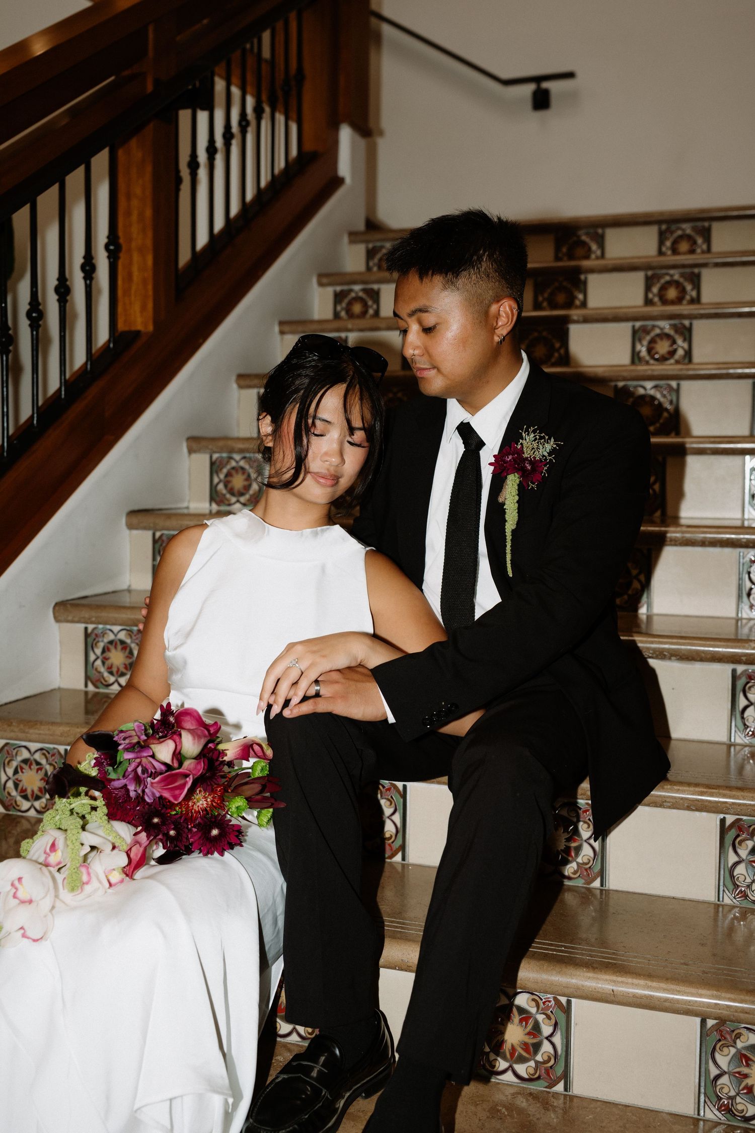 A bride and groom sit together on tiled stairs, she holds a burgundy bouquet as he gazes at her lovingly.