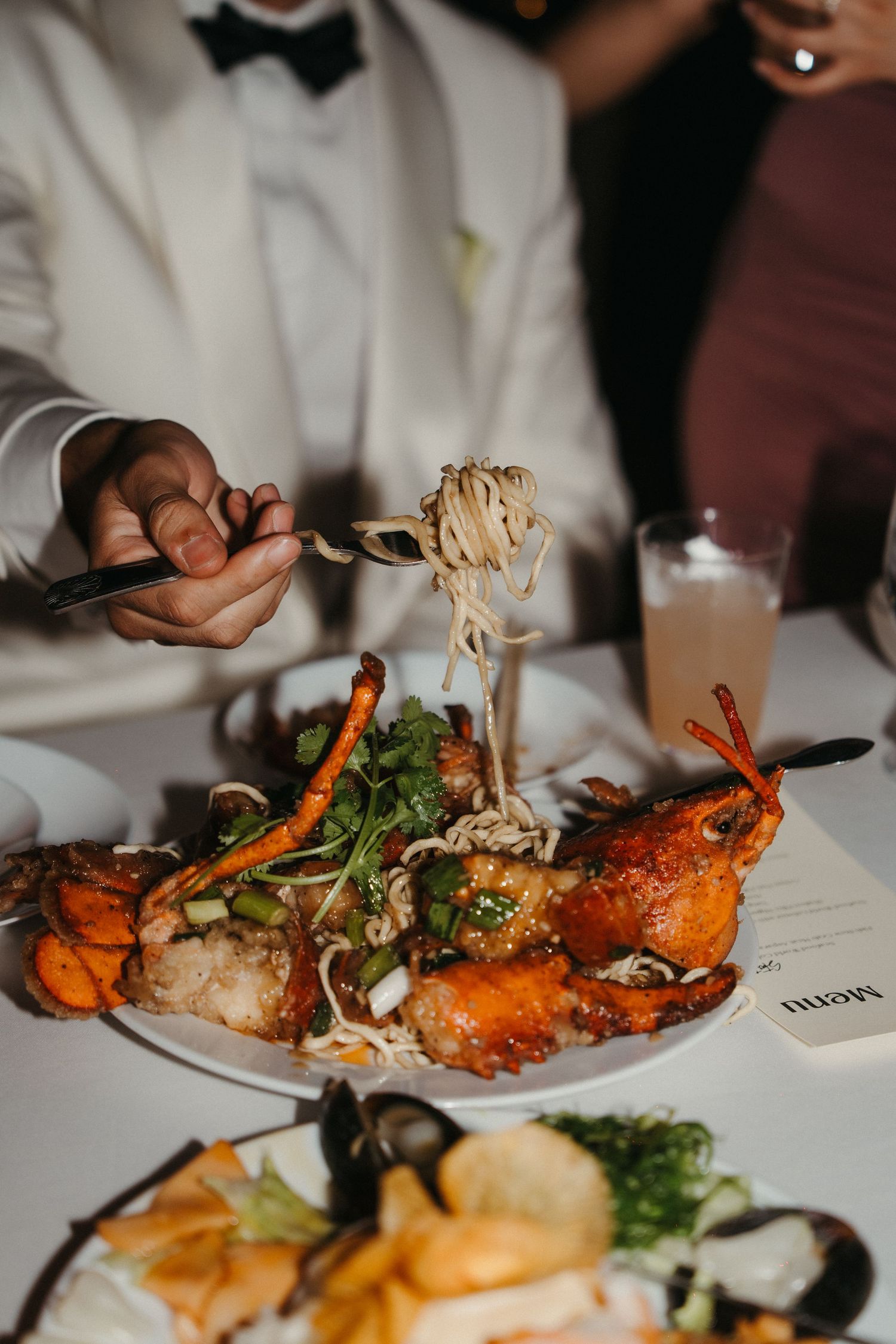 Groom lifting noodles with fork over a seafood platter with lobster and greens during wedding reception dinner.