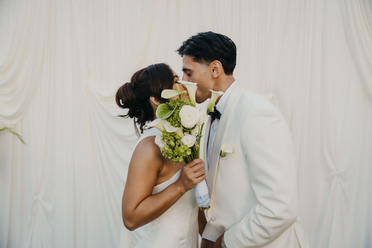 Bride and groom sharing a kiss behind a white floral bouquet at an elegant outdoor wedding cocktail hour.