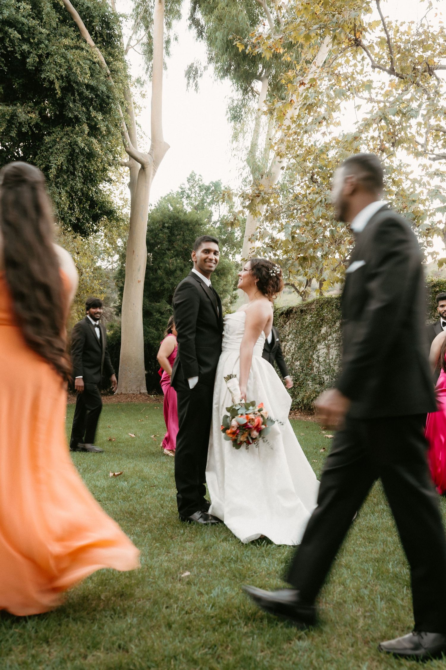 Bride and groom share a moment on green lawn surrounded by bridal party on their wedding day.
