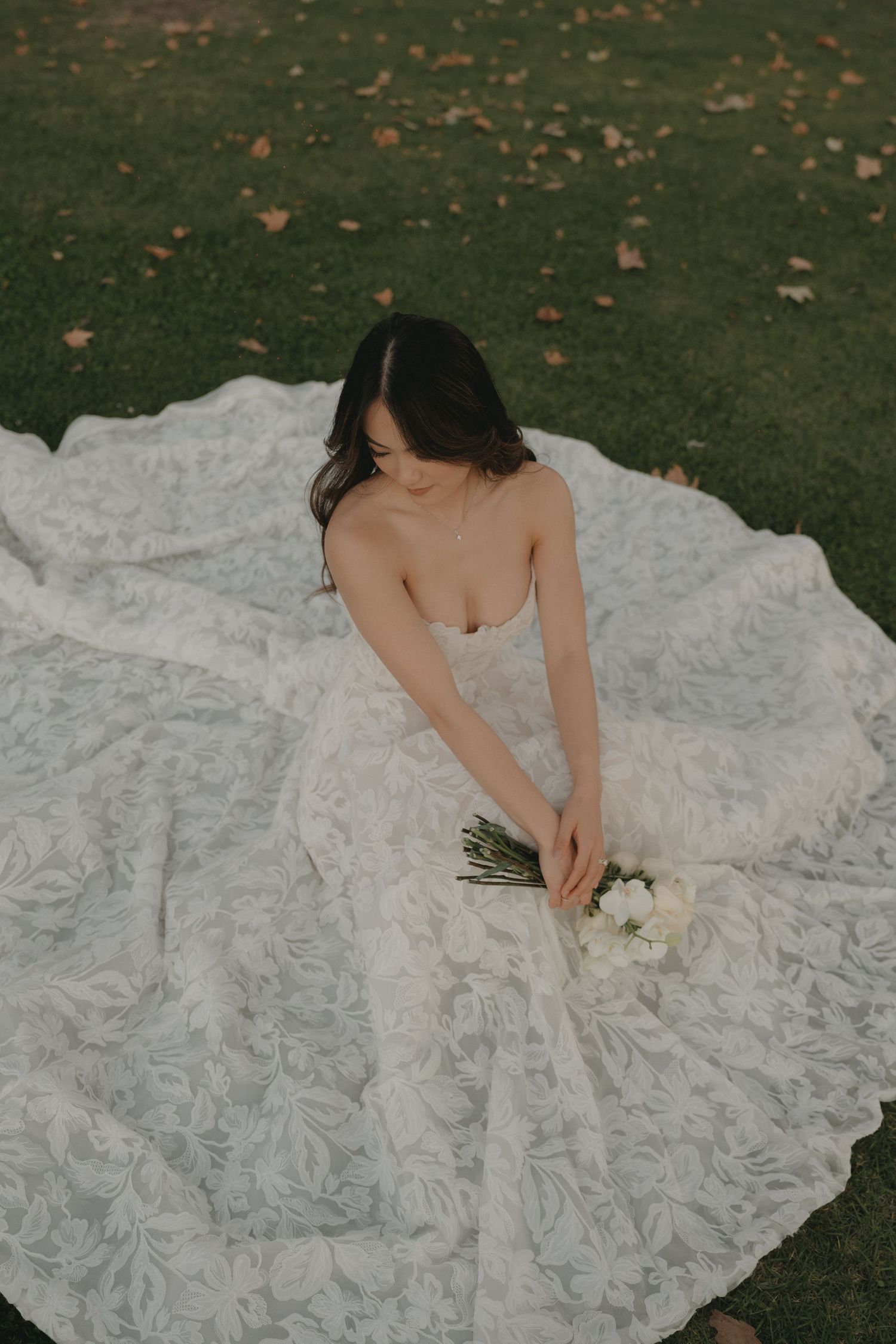 Bride sitting on grass holding a bouquet on her wedding day.