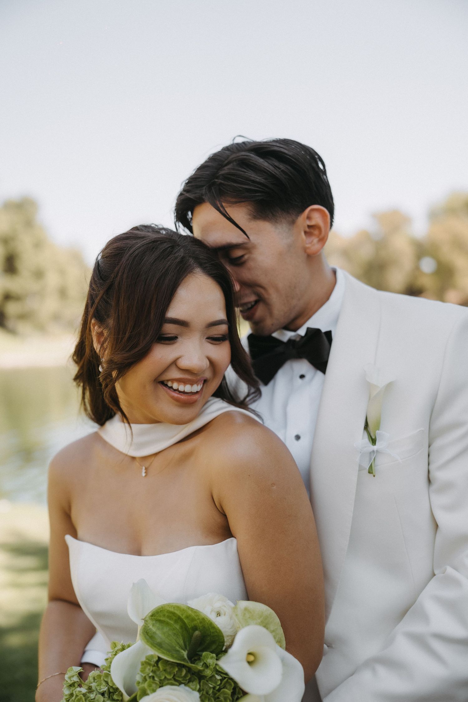 Smiling bride in strapless white gown holds calla lily bouquet as groom in white tuxedo nuzzles her cheek outdoors.