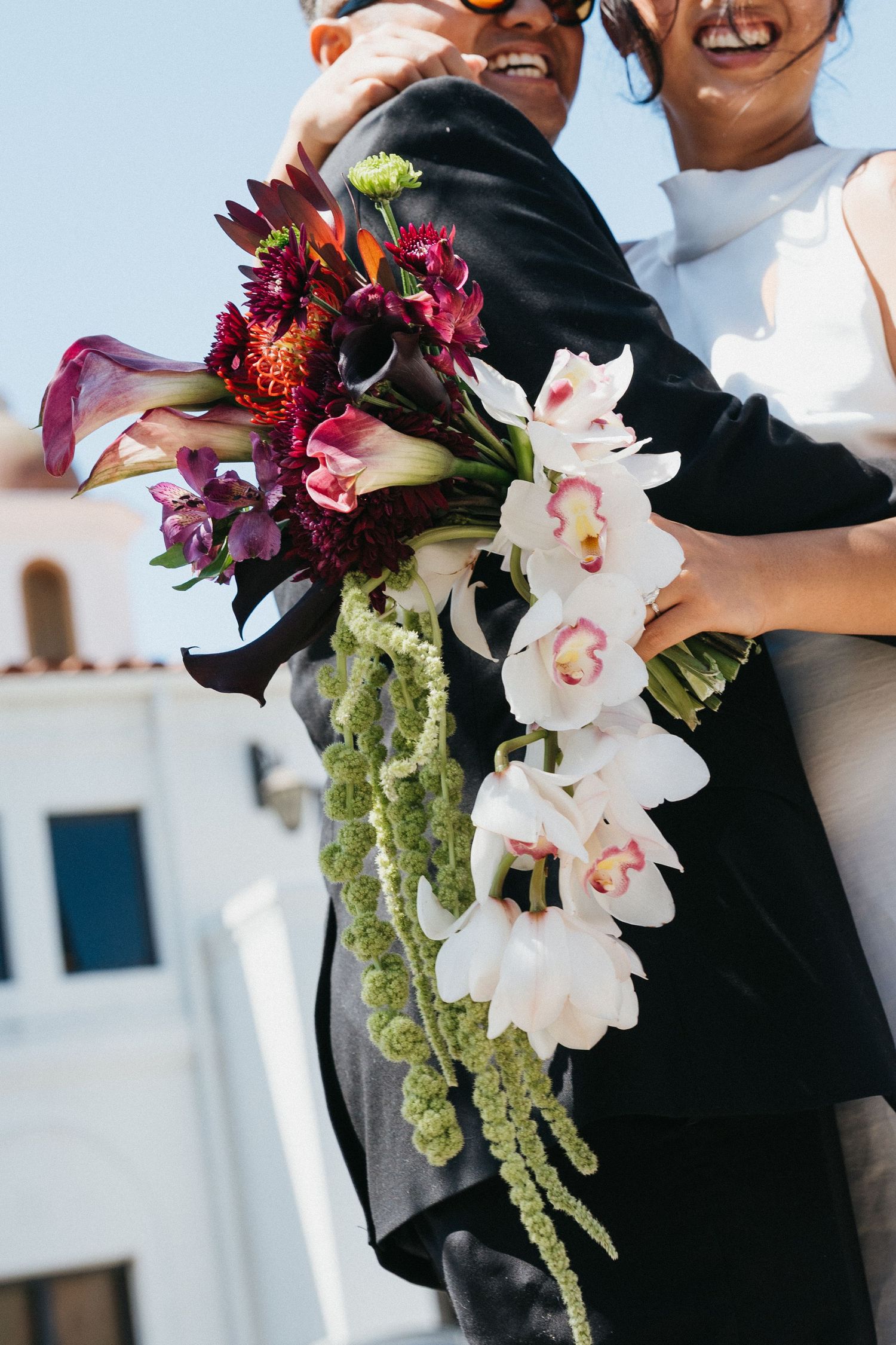 Image of bridal bouquet with Orchids and burgundy flowers while newlyweds smile and the sky in background.