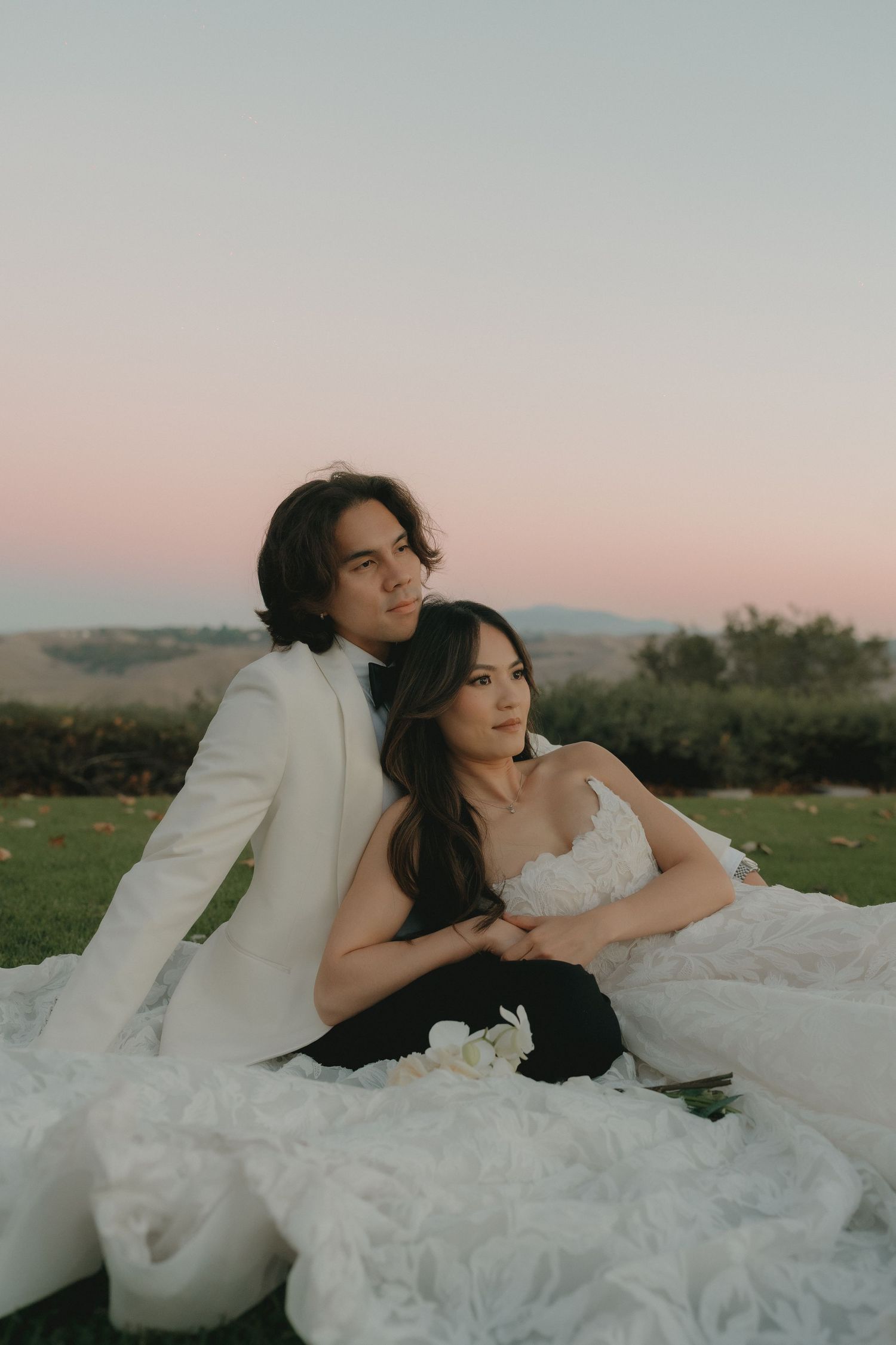 Newlyweds sitting on grass at sunset, the groom in white suit embracing the bride in a flowing white gown.