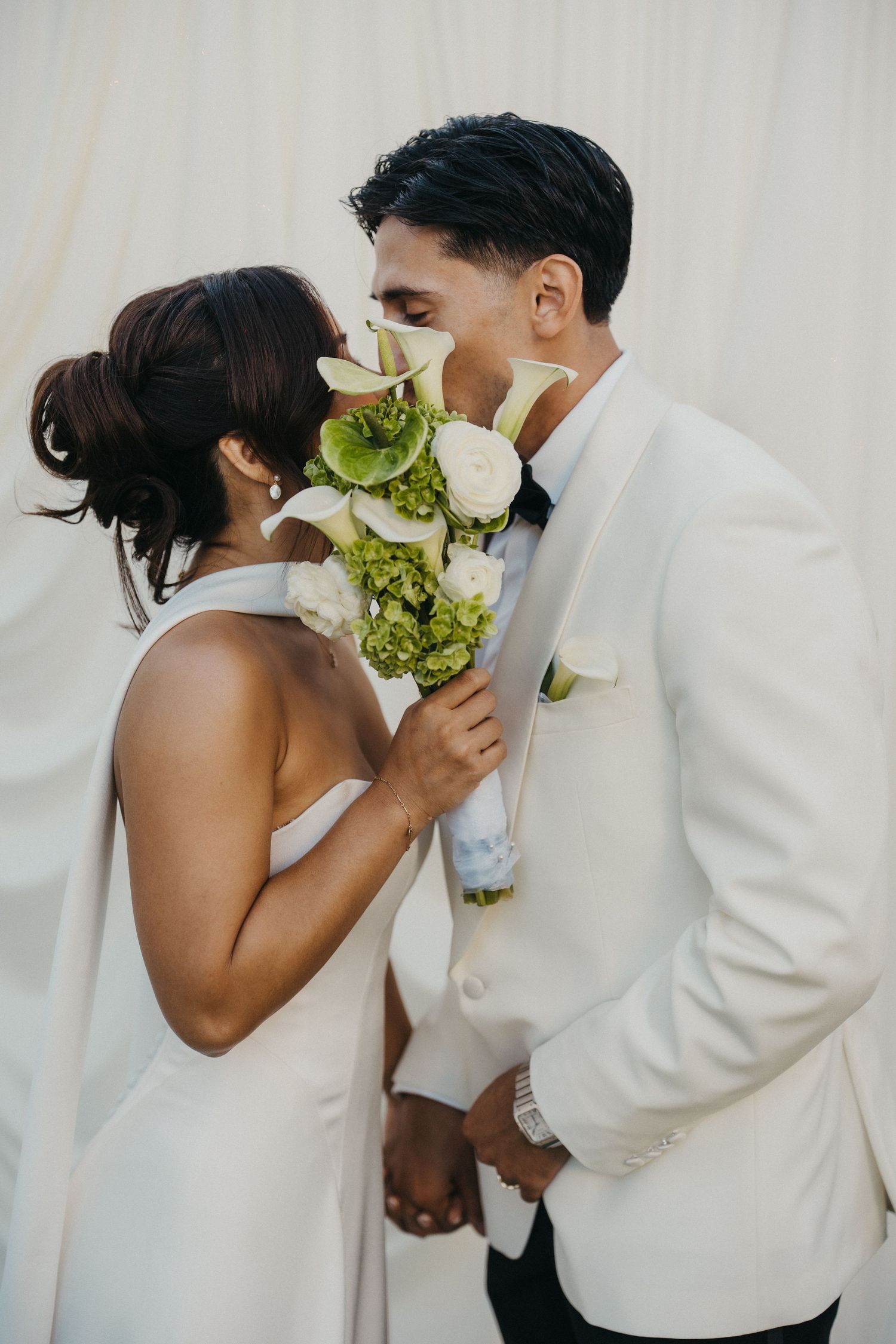 Bride and groom share a kiss behind a white and green floral bouquet at their elegant outdoor wedding cocktail hour.