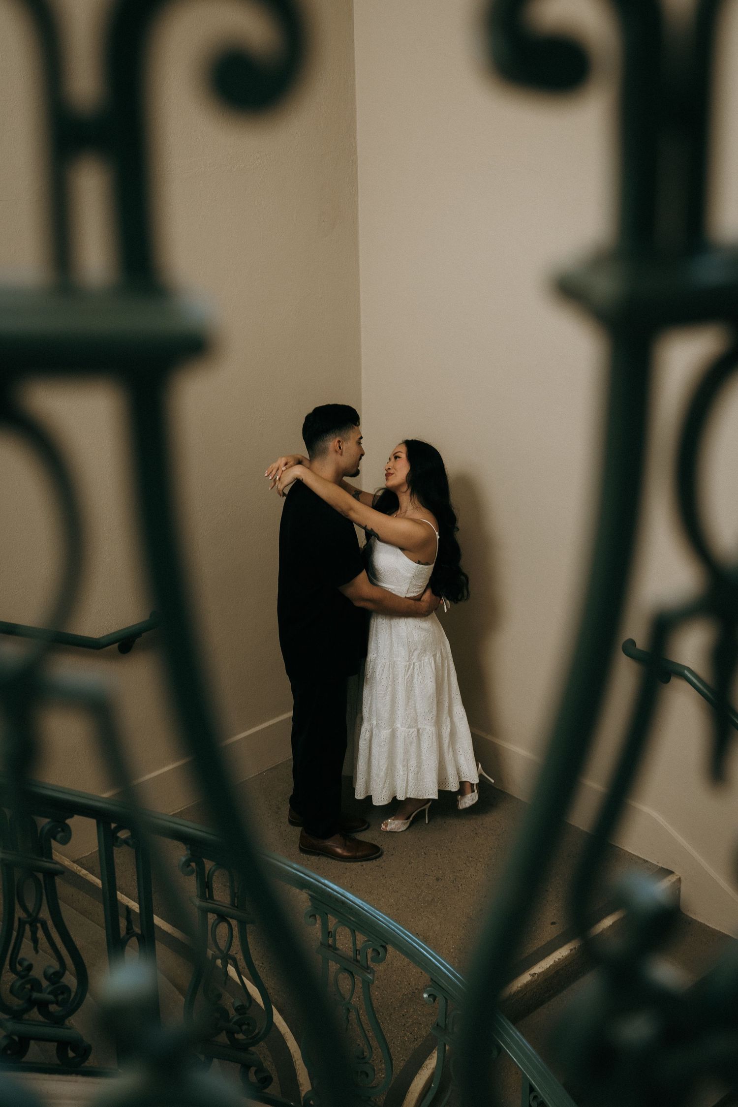 Couple embracing in Pasadena City Hall stairwell, shot from afar with railing in the foreground.