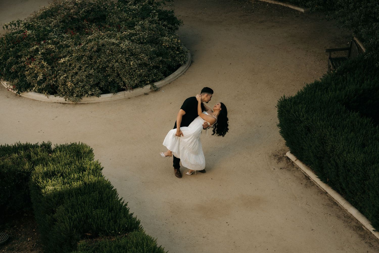 Couple sharing a romantic dip dance pose on a garden path surrounded by lush greenery, aerial view.