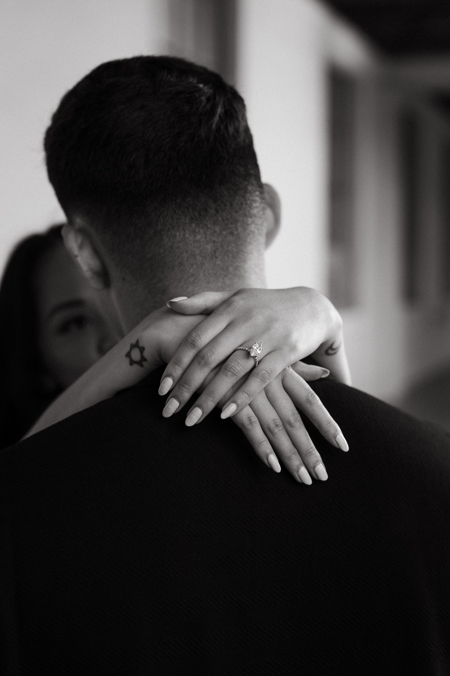 Black and white photo of a couple embracing, womans hand with engagement ring resting on mans shoulder.