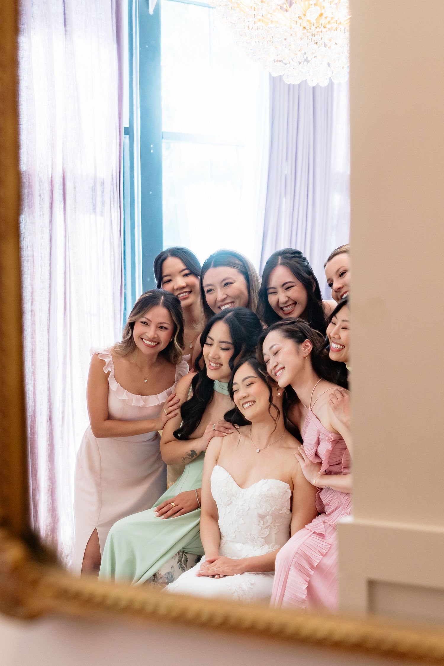 Bride in white gown sits surrounded by smiling bridesmaids in pastel dresses, reflected in a gold-framed mirror.