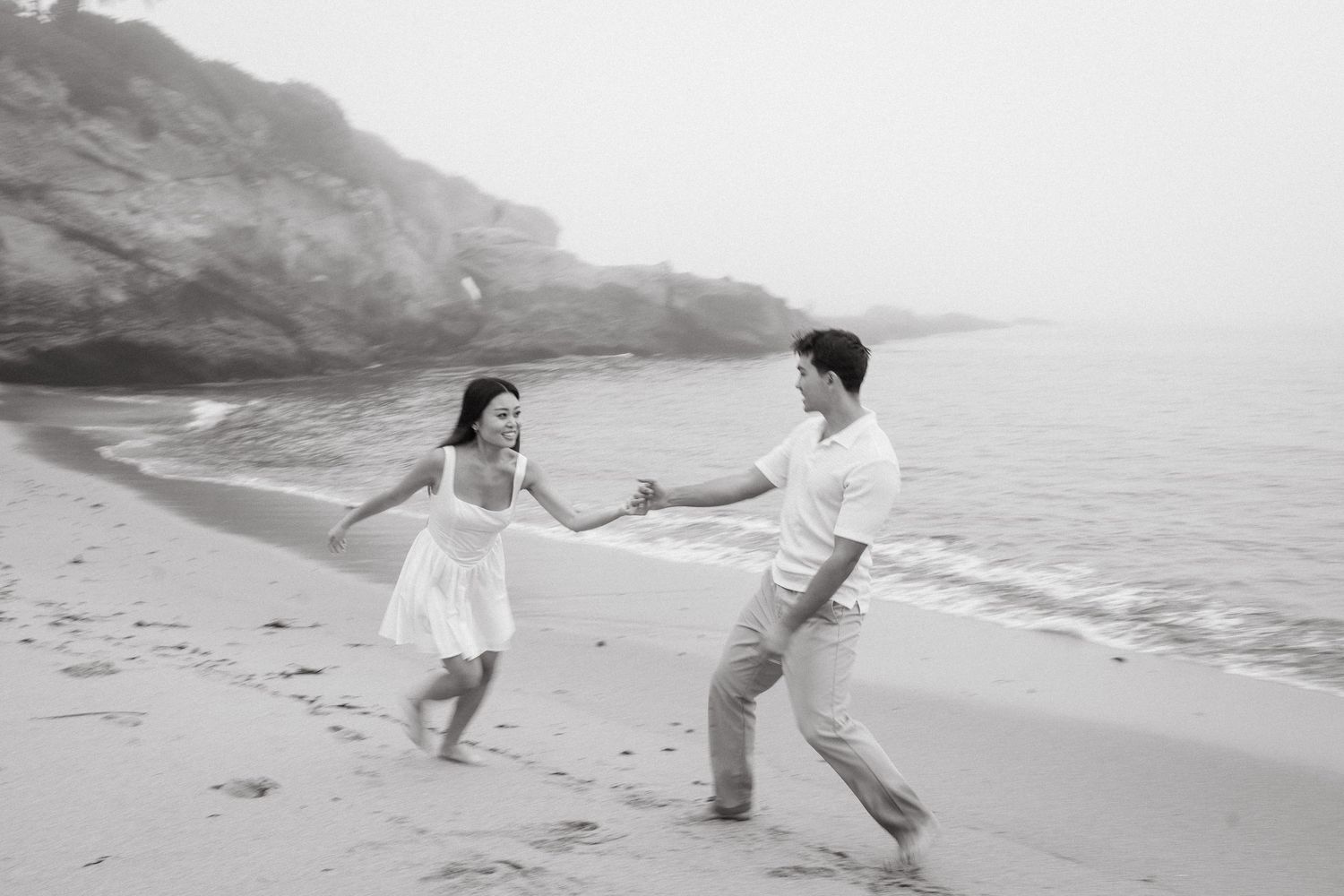 A couple joyfully holds hands and dances on a misty beach, with rocky cliffs in the background.