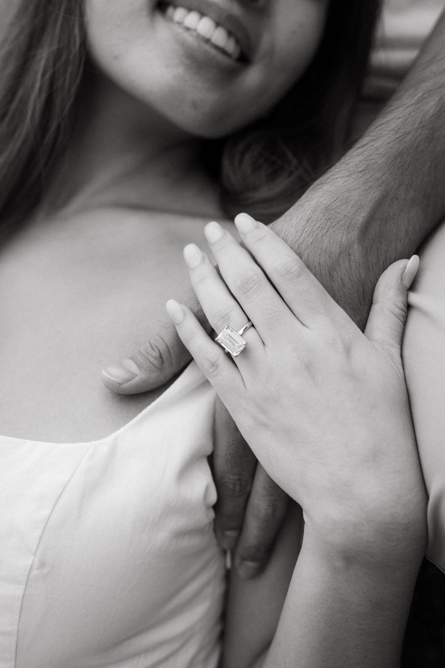 Black and white photo of a smiling bride showing her elegant emerald cut diamond engagement ring while holding her partner's arm.