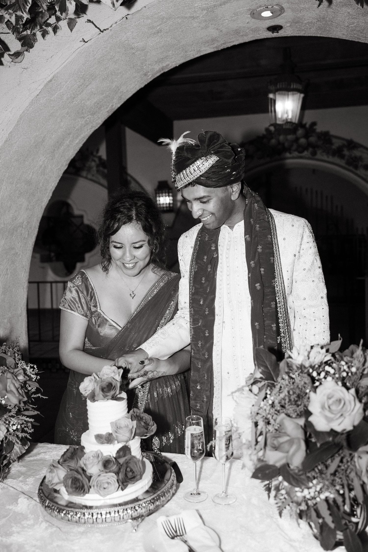 Bride and groom in traditional Indian attire cutting wedding cake under stone arch in black and white photo.