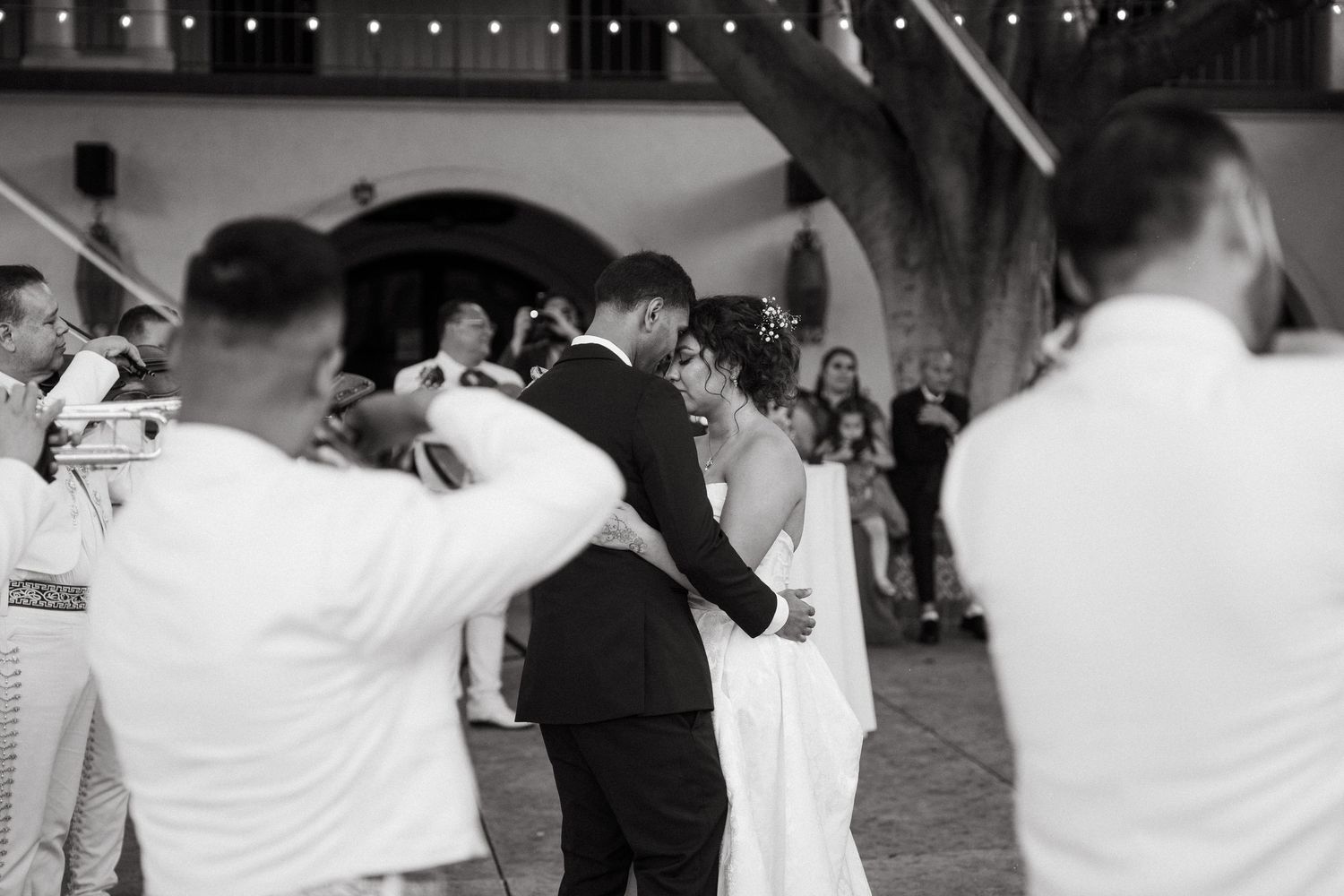 Black and white photo of a bride and groom sharing their first dance surrounded by mariachi musicians at their wedding.
