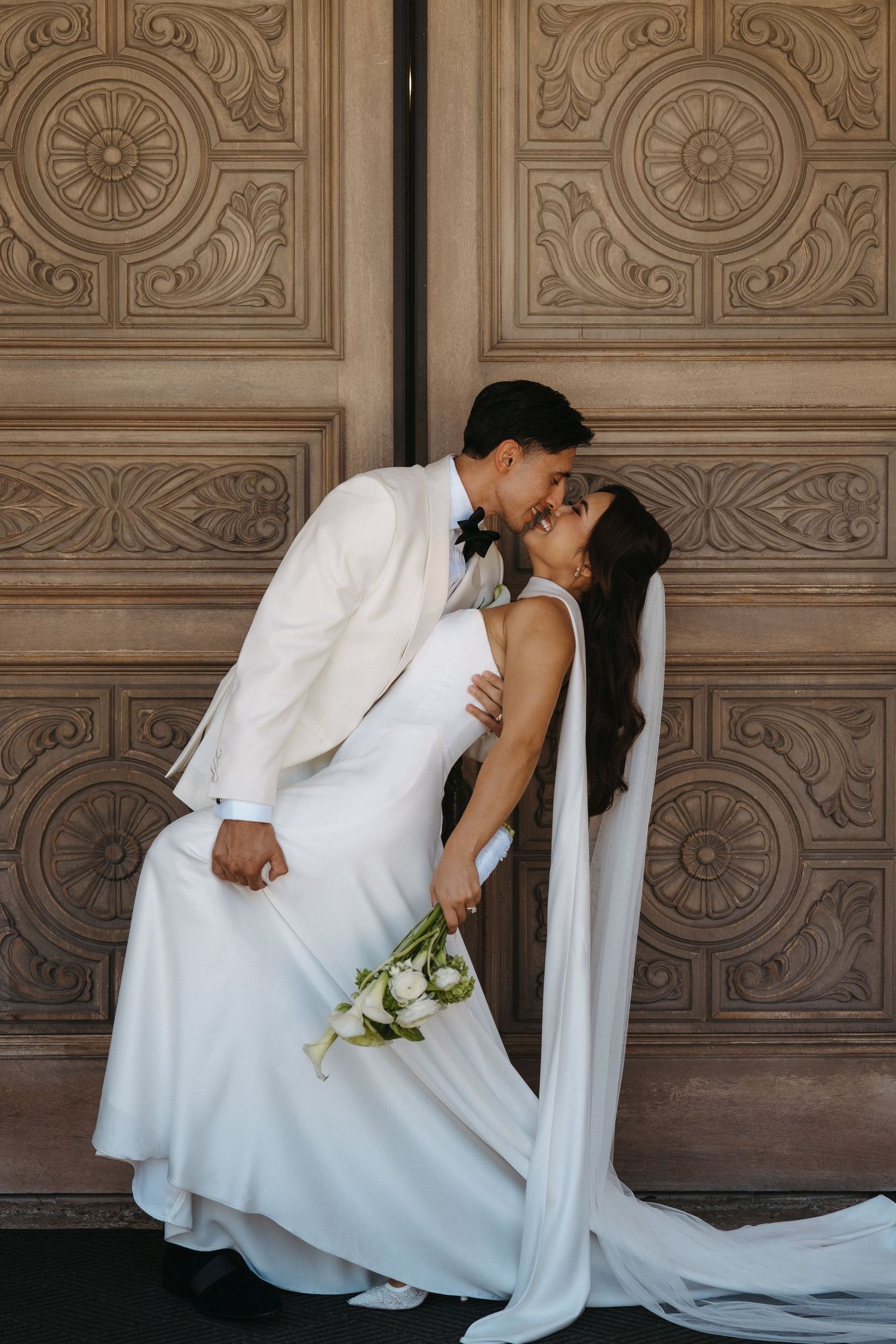 Bride and groom share a romantic kiss in front of church doors, bride holding white bouquet.