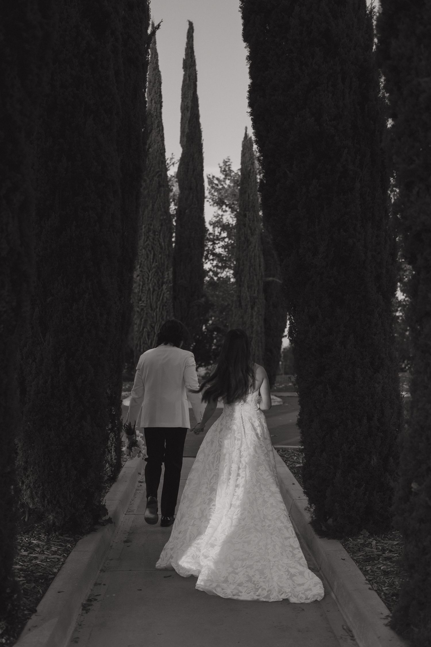 Black and white image of newlyweds walking through a path of trees with dramatic shadows.