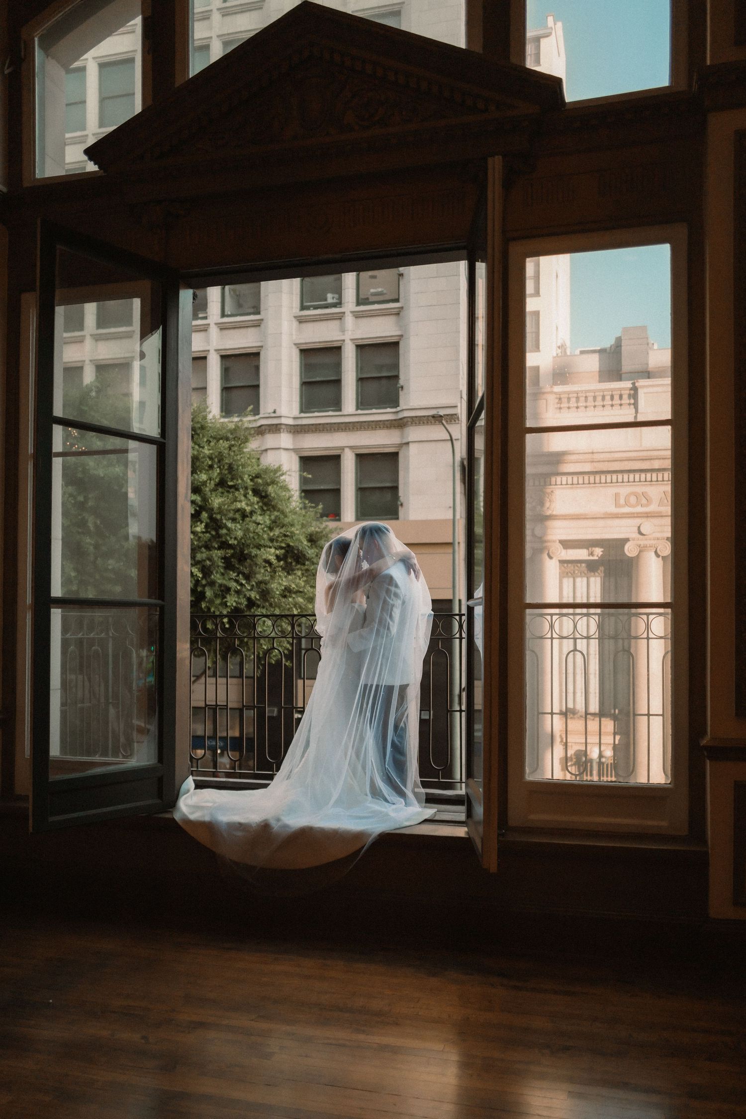 Newlyweds embracing under bride's veil while standing in the opening of a large window on their wedding day.