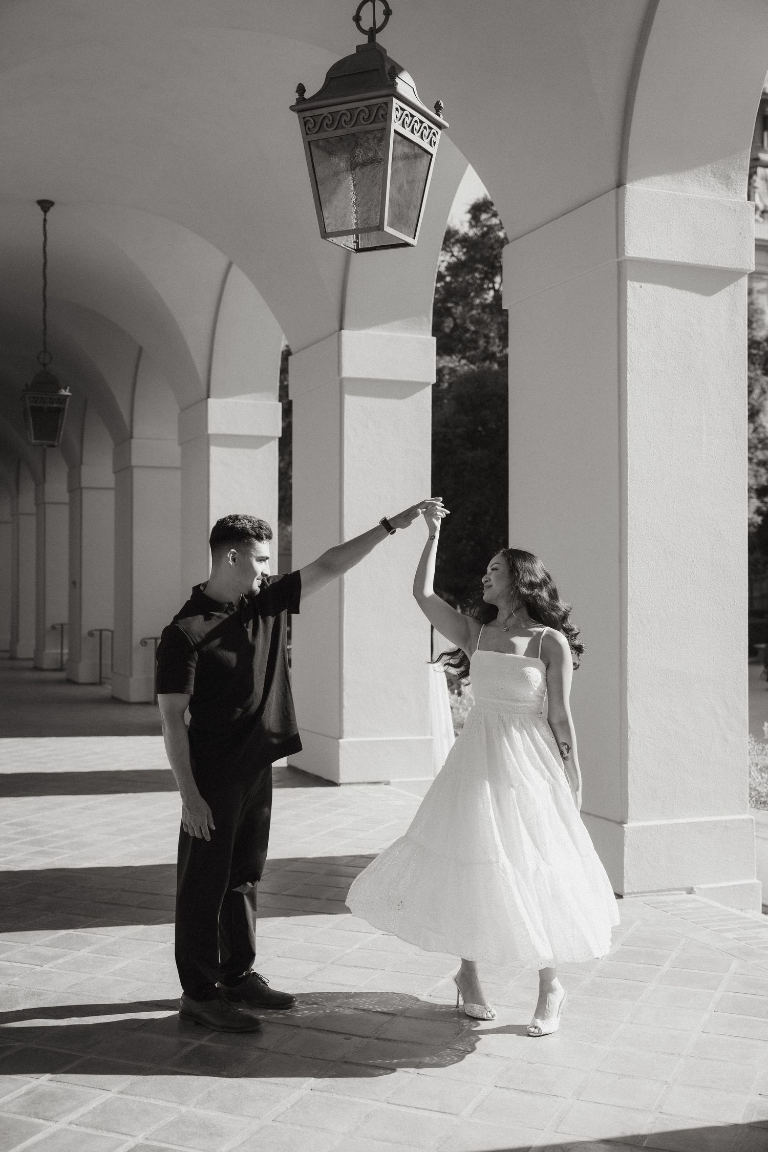 Black and white photo of a couple dancing under arched colonnade, woman in white dress being twirled by man in black.