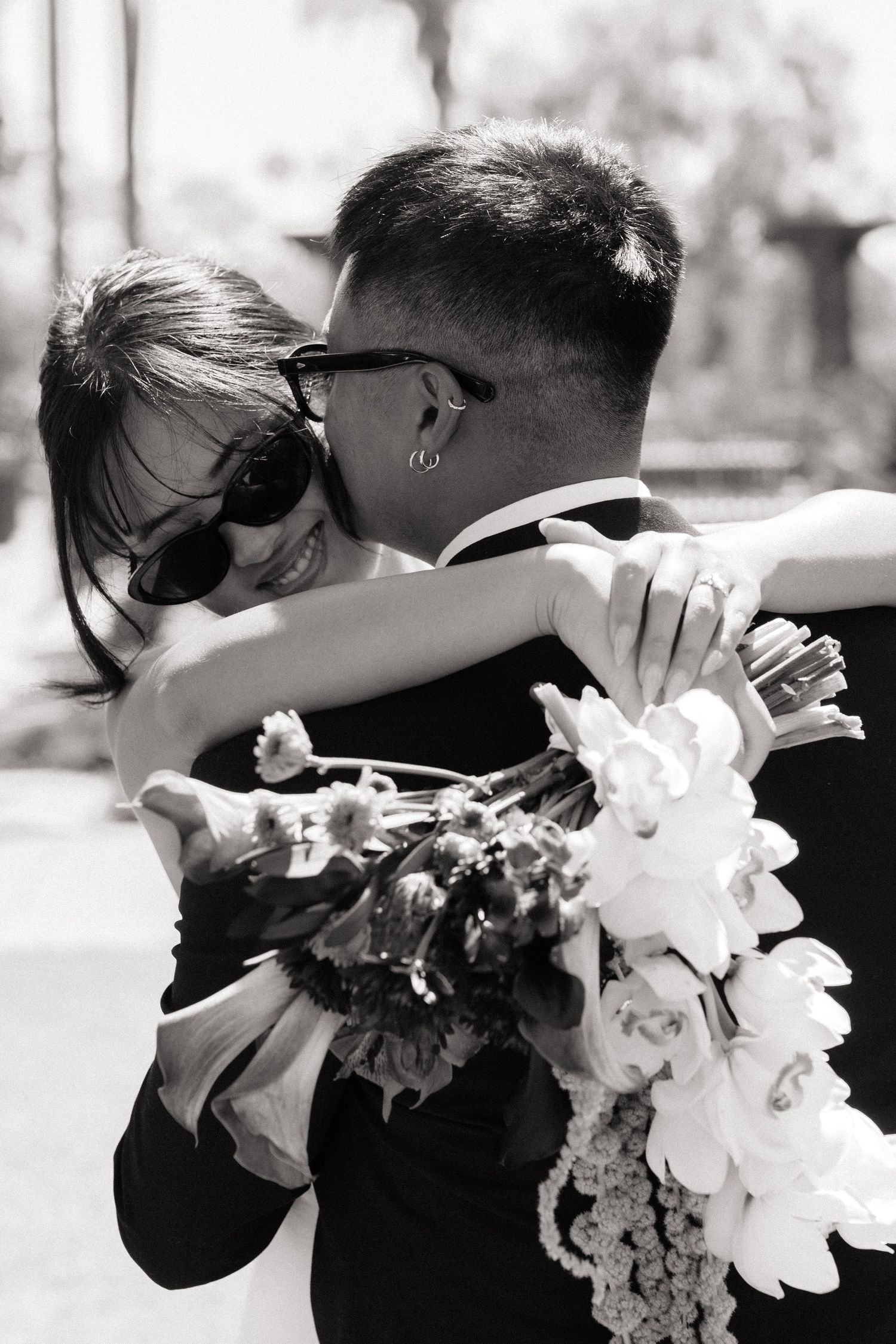 Black and white photo of a smiling bride hugging groom while holding a floral bouquet, both wearing sunglasses outdoors.