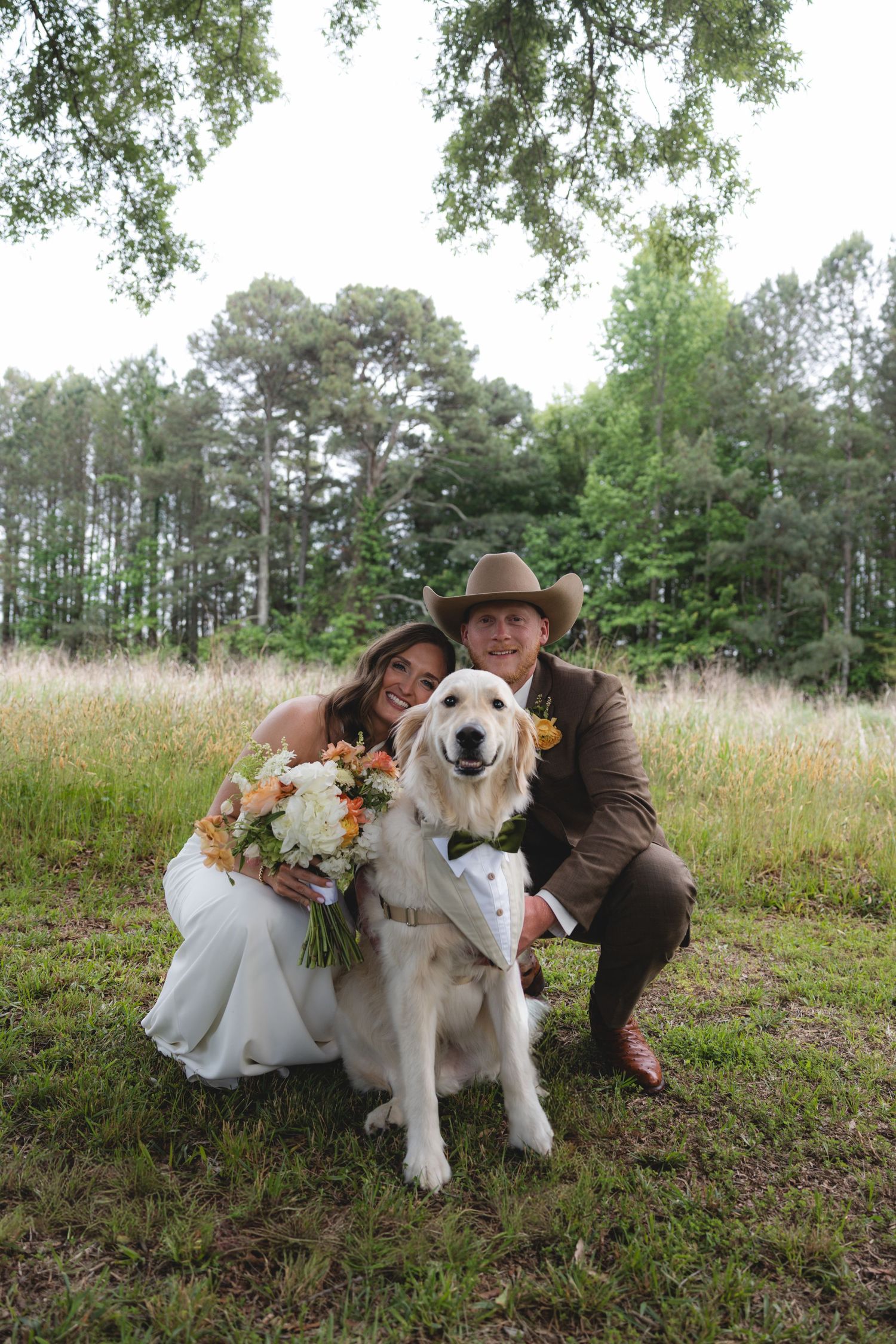 Bride and groom pose with their golden retriever wearing a tuxedo bandana at an outdoor rustic wedding ceremony.