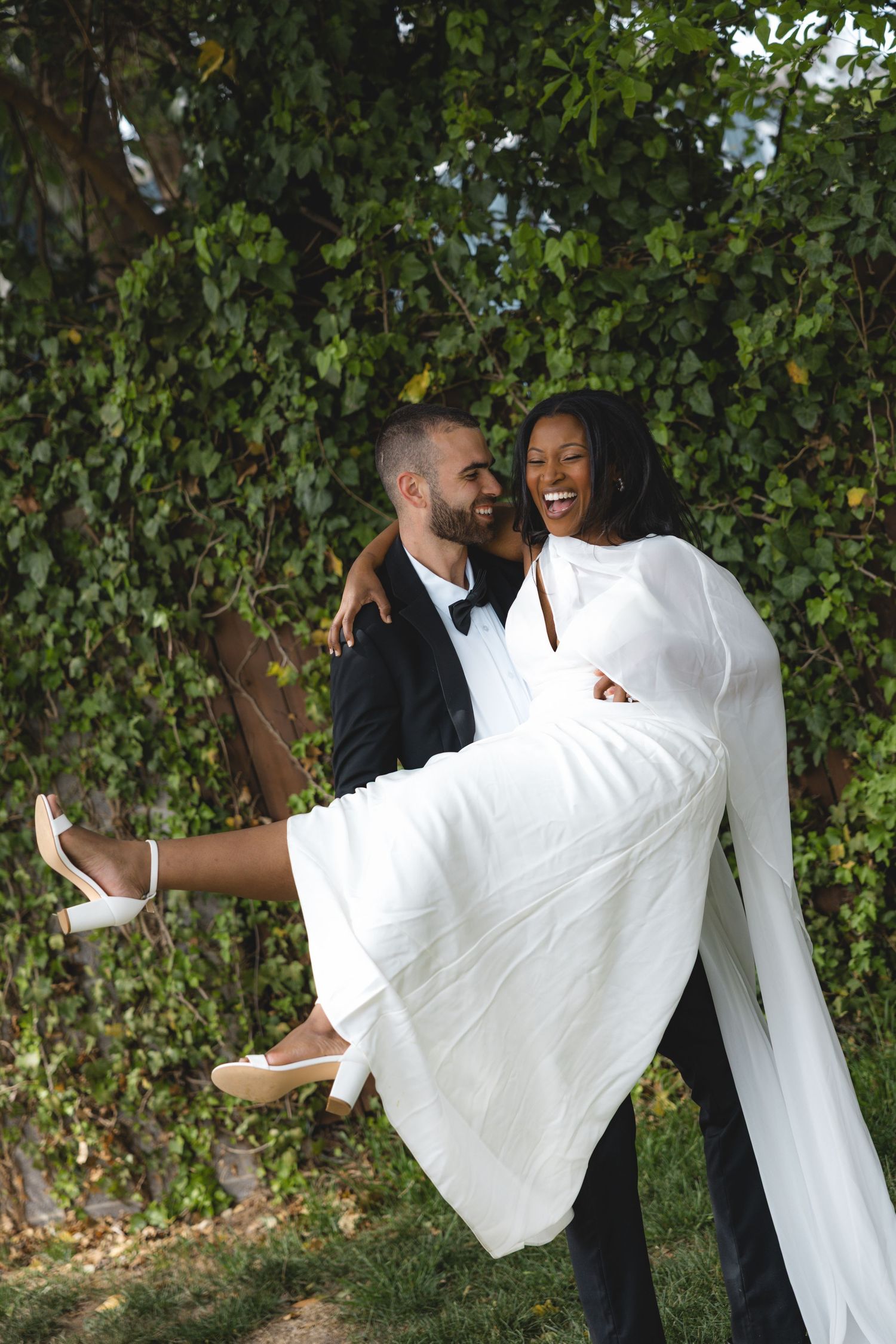 Groom in tuxedo carries smiling bride in white wedding dress and cape outdoors near lush green ivy wall.