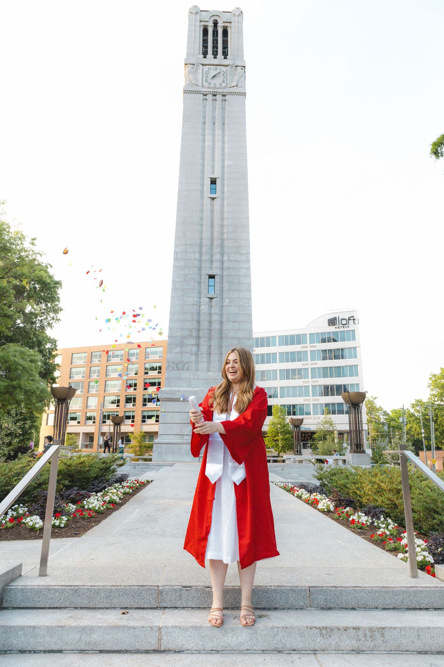 Young woman in red graduation gown stands proudly on steps before a tall bell tower on a bright day.