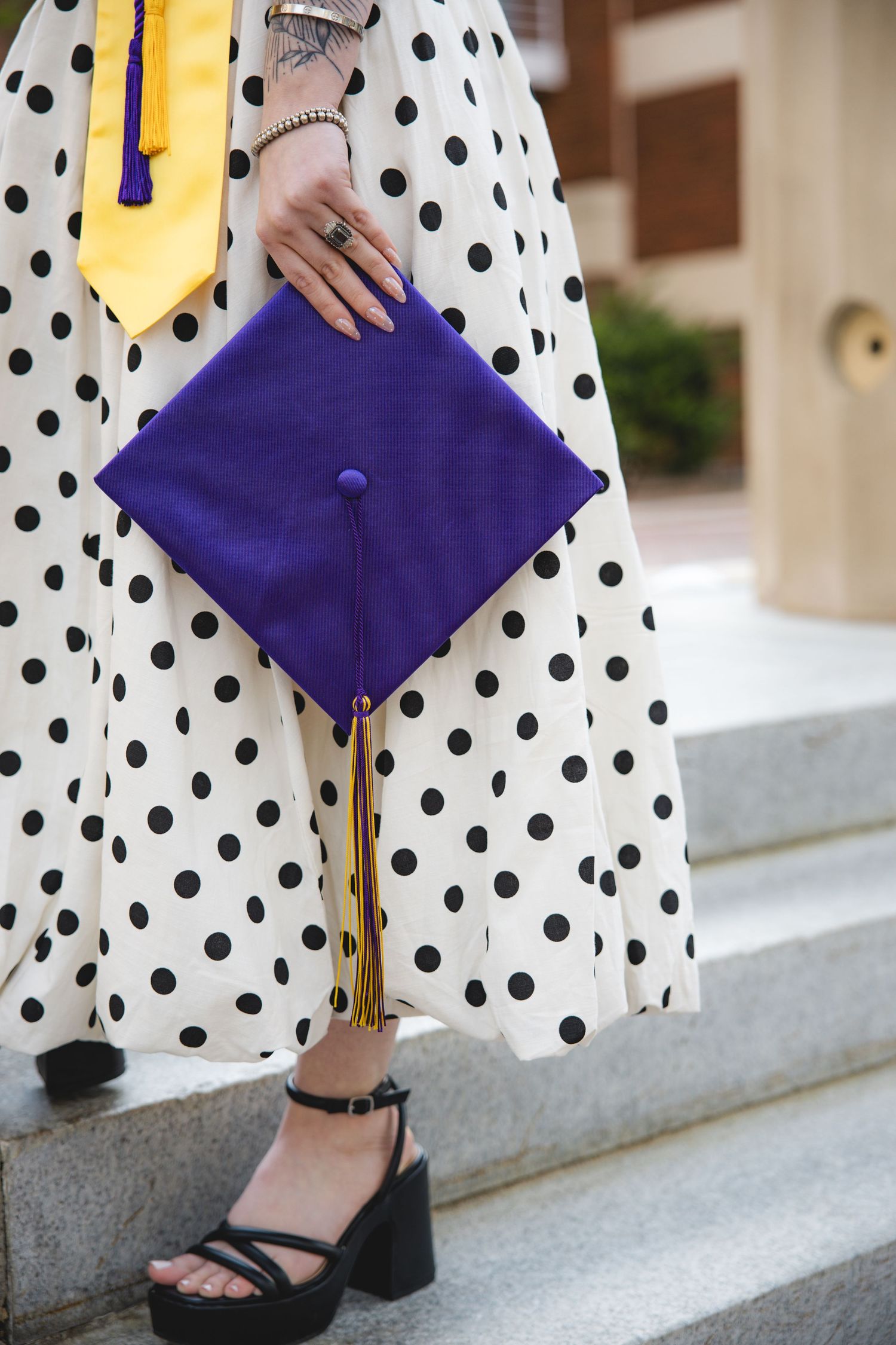 Graduate holding purple mortarboard cap with gold tassel, wearing polka dot dress and black heeled sandals on stone steps.
