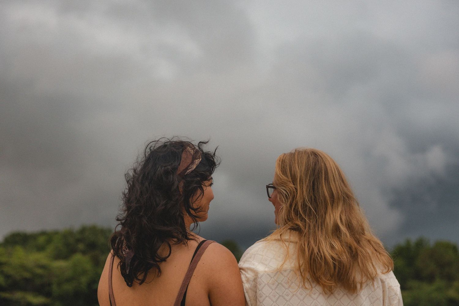 Two women with long hair stand together outdoors, gazing at a dramatic stormy sky above lush green trees.