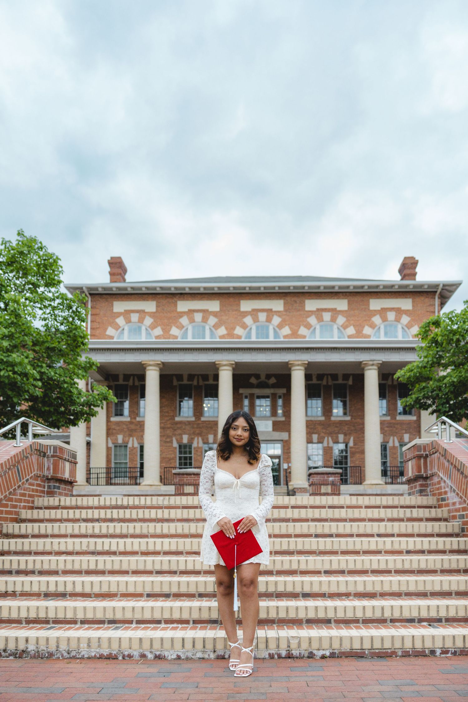 Woman in white lace dress holding red clutch stands on brick steps before a classic colonial brick building.