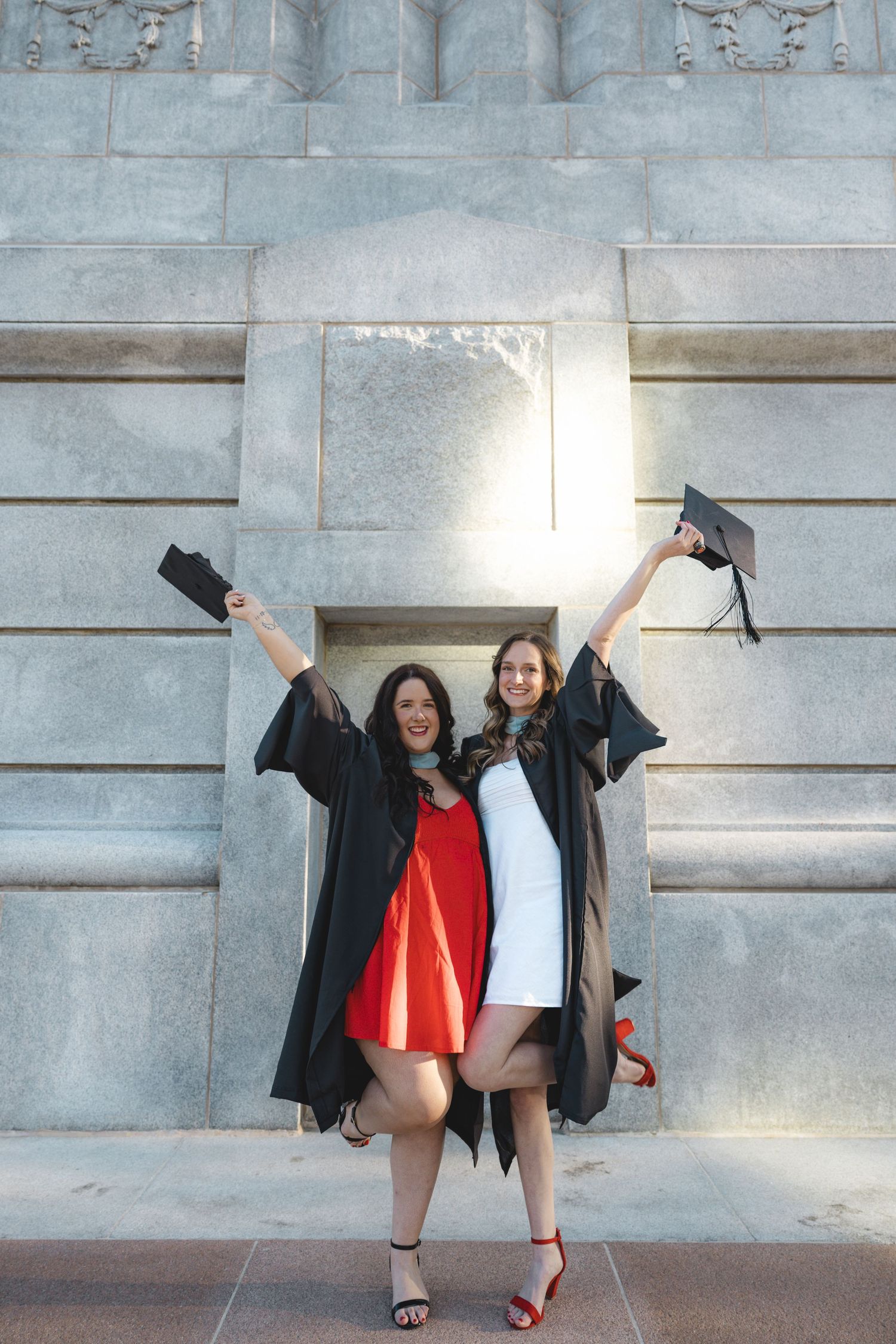 Two happy graduates in black gowns celebrate outdoors, raising their caps while wearing red and white dresses.