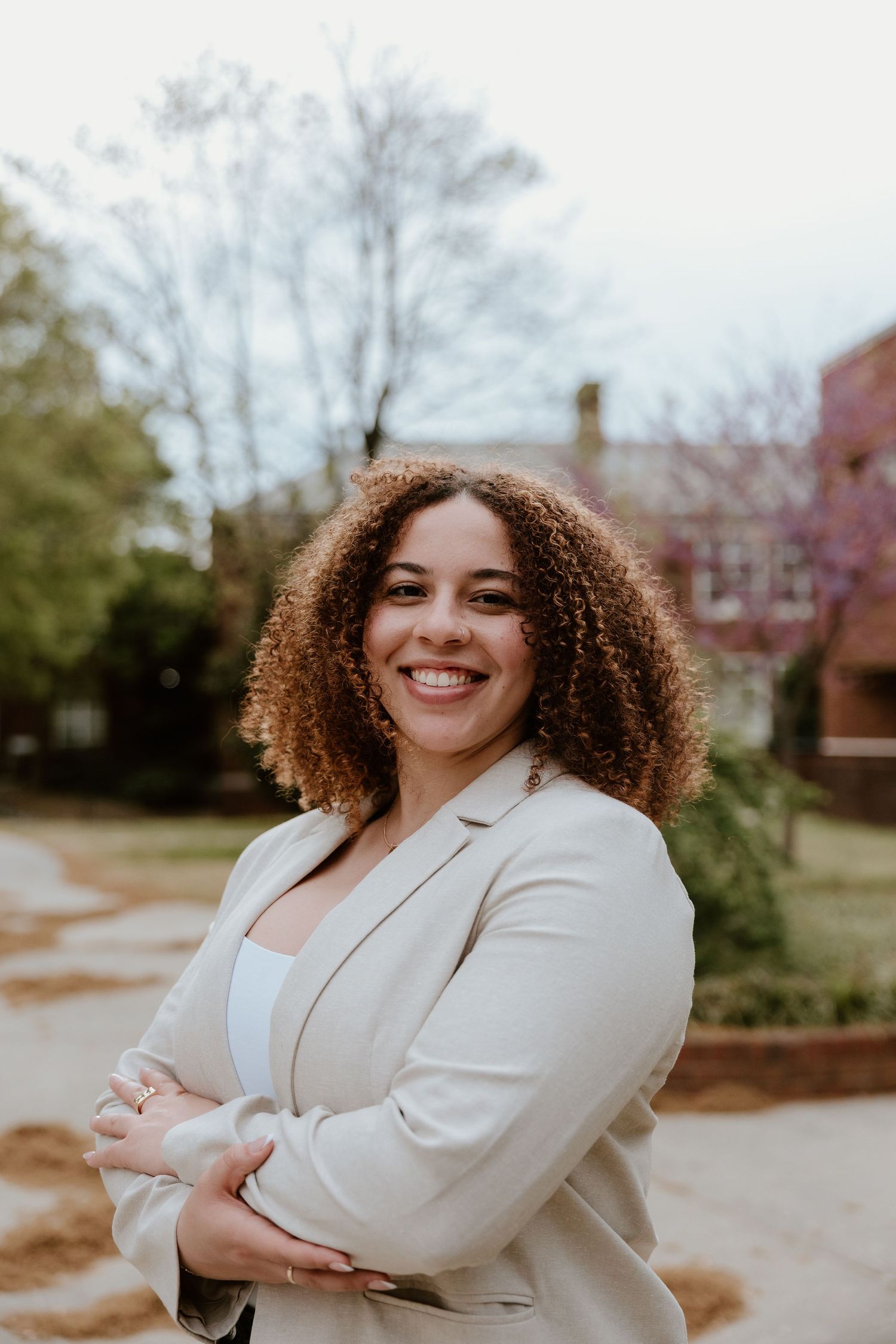 Smiling young woman with curly hair in a beige blazer, arms crossed, posing outdoors near brick buildings.